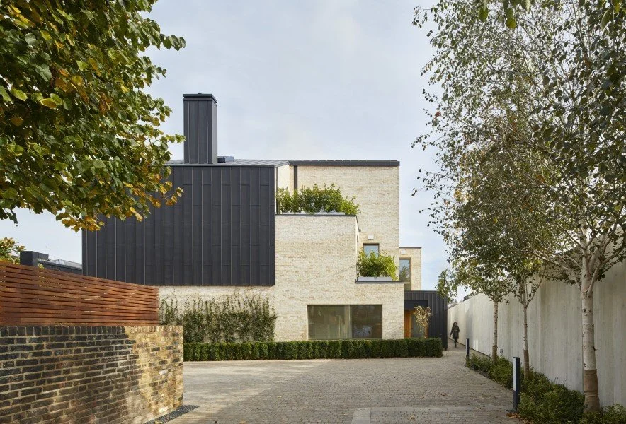 A modern multi-story building with a combination of black and light-colored brick exterior, large windows, and balconies with plants, situated along a paved pathway with trees and a white wall.