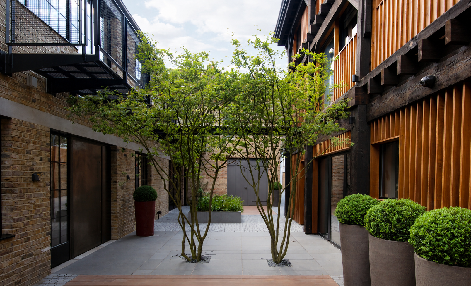 A modern outdoor courtyard with brick and wood siding buildings, a small tree in the center, potted plants, and a sky with some clouds overhead.