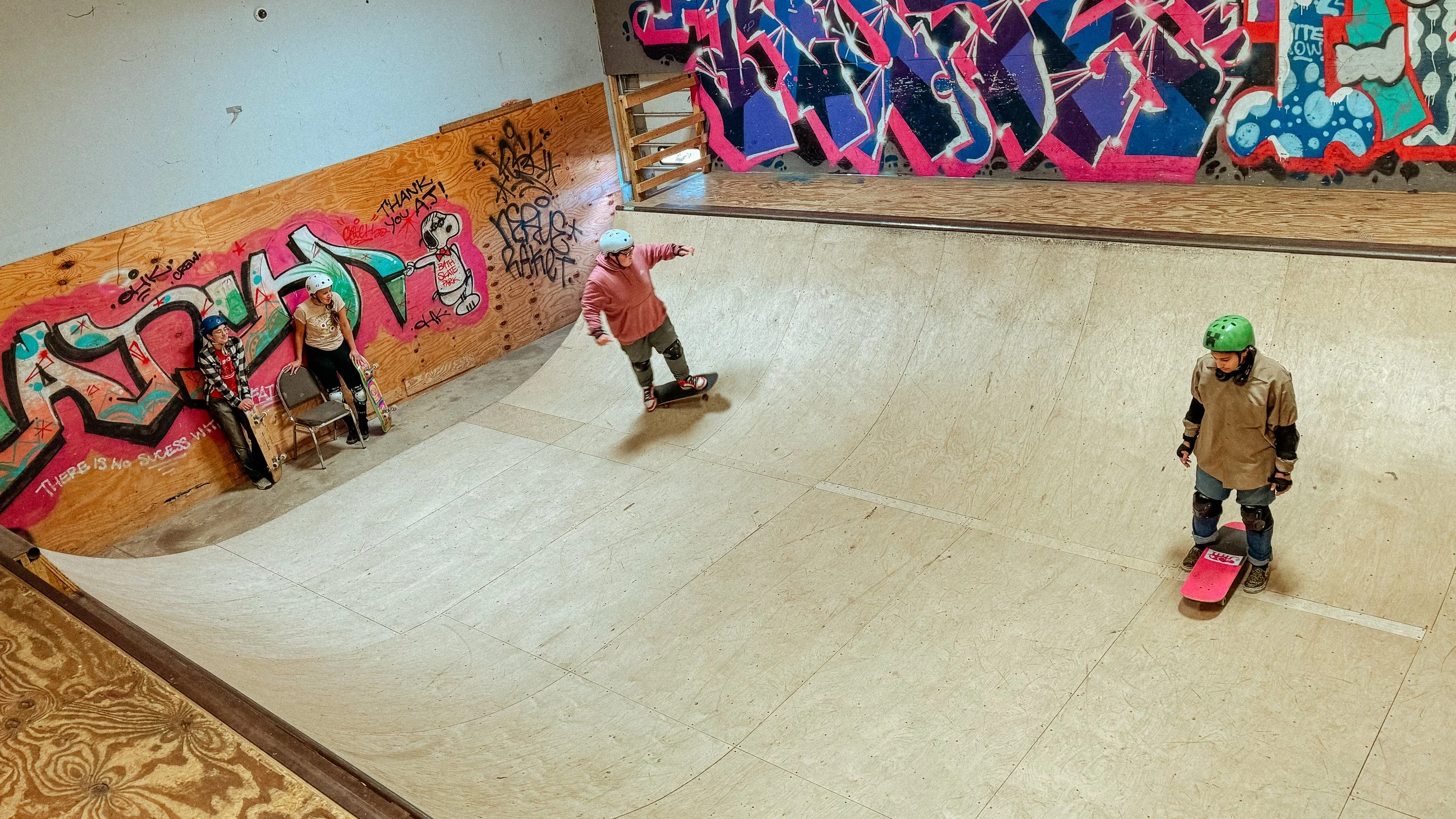 image from above of a young person skating a wooden ramp, with two people watching. it's indoor with graffitti.