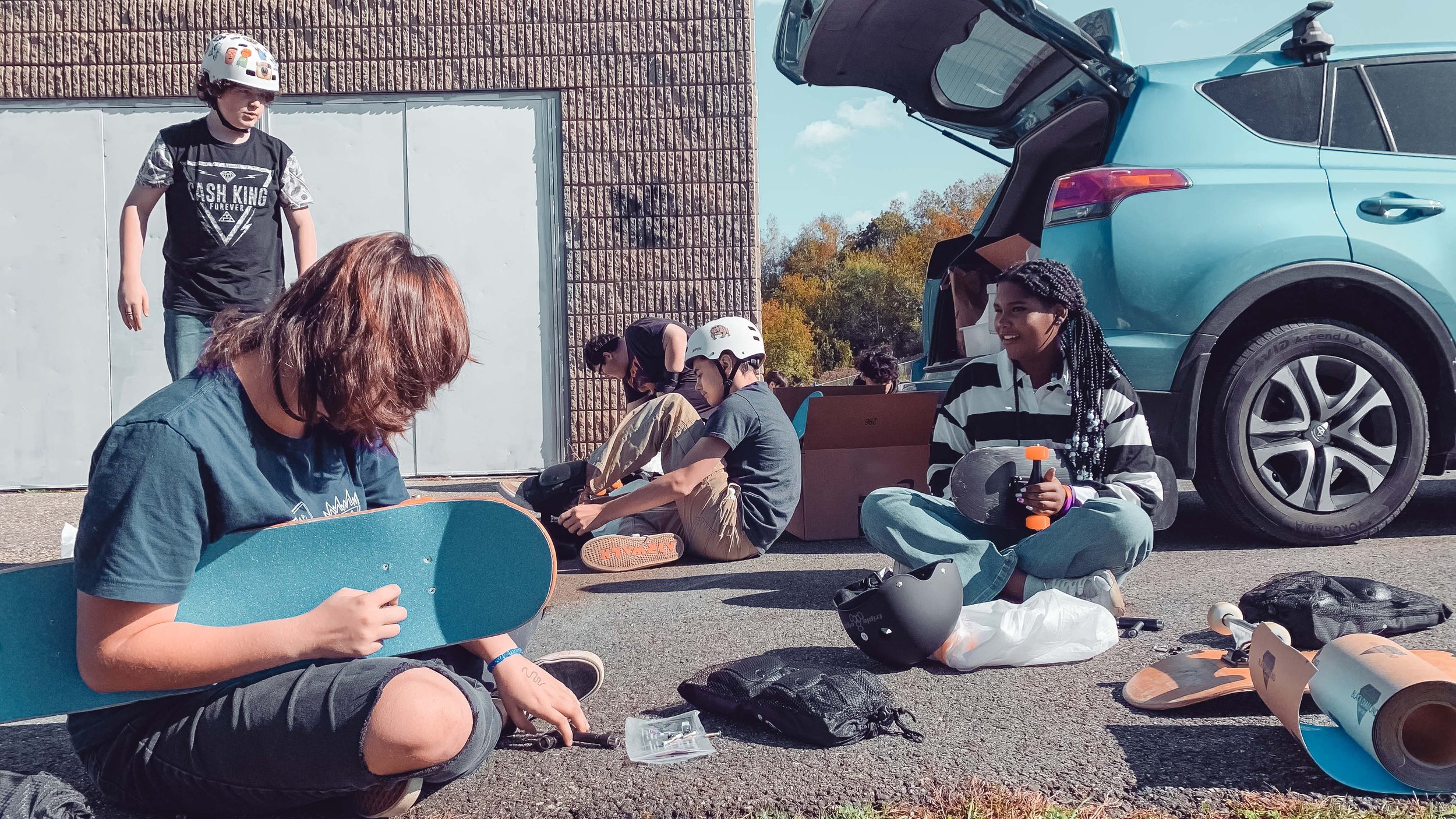 youth in the program sitting in a parking lot, putting shoes on, pads on, and adjusting skateboards.