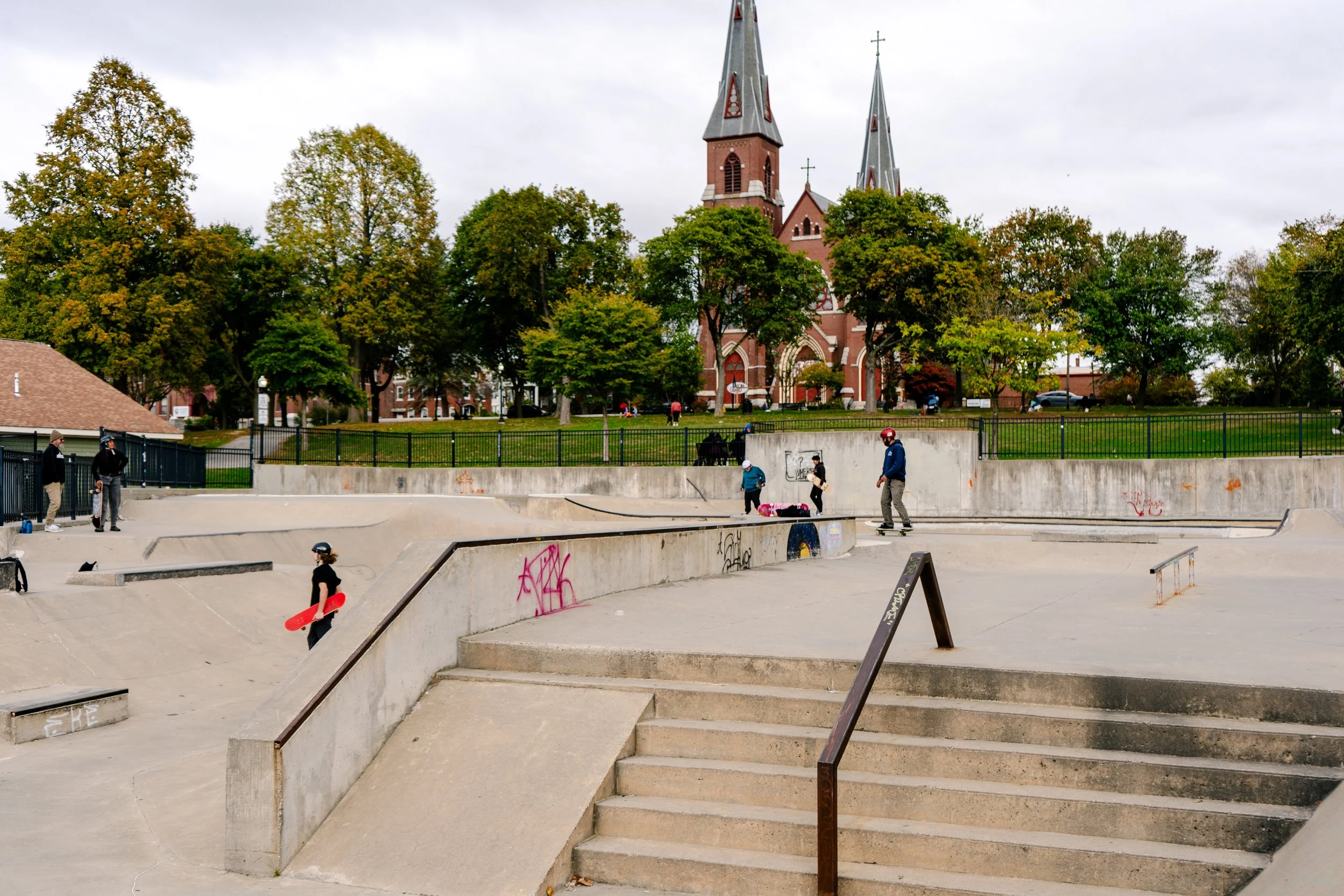 far away landscape style shot of a skatepark, backed by a regular park. people are skating and hanging out.
