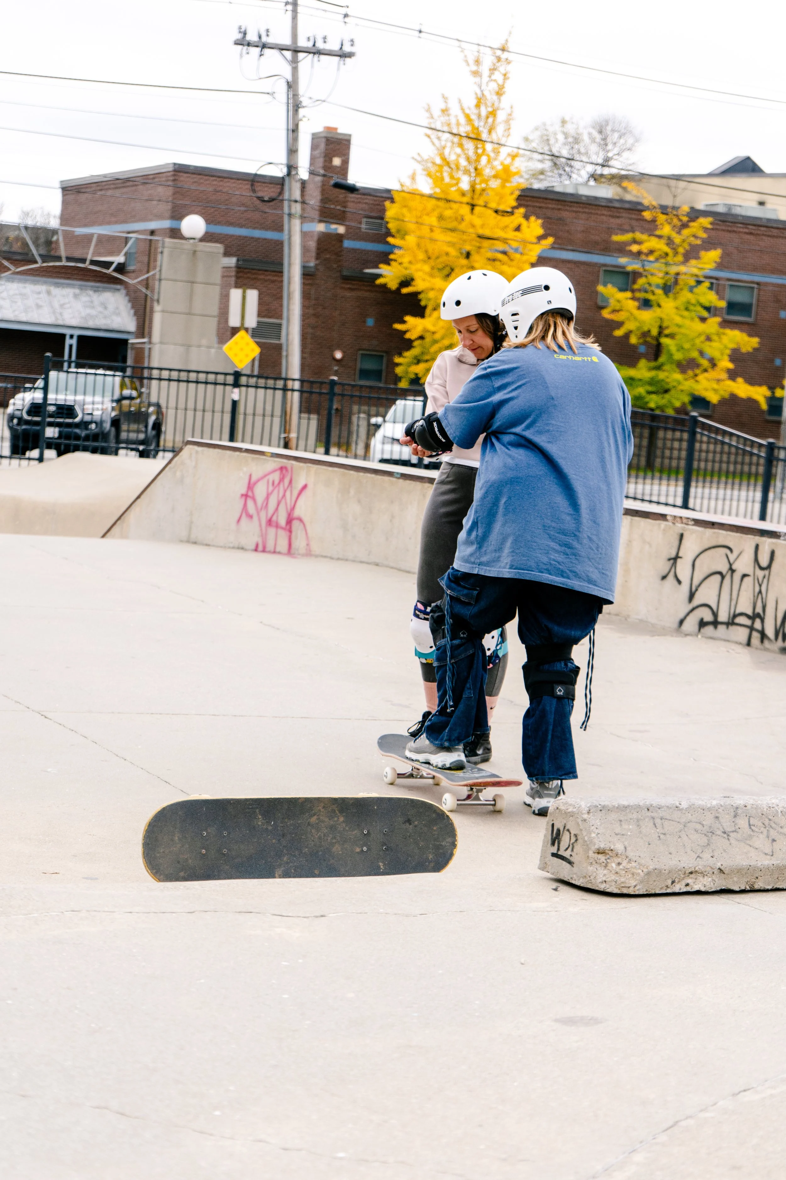 Two skateboarders wearing helmets and protective gear at a skate park, with one standing on a skateboard and the other helping. Graffiti and a concrete barrier are visible, with autumn trees in the background.