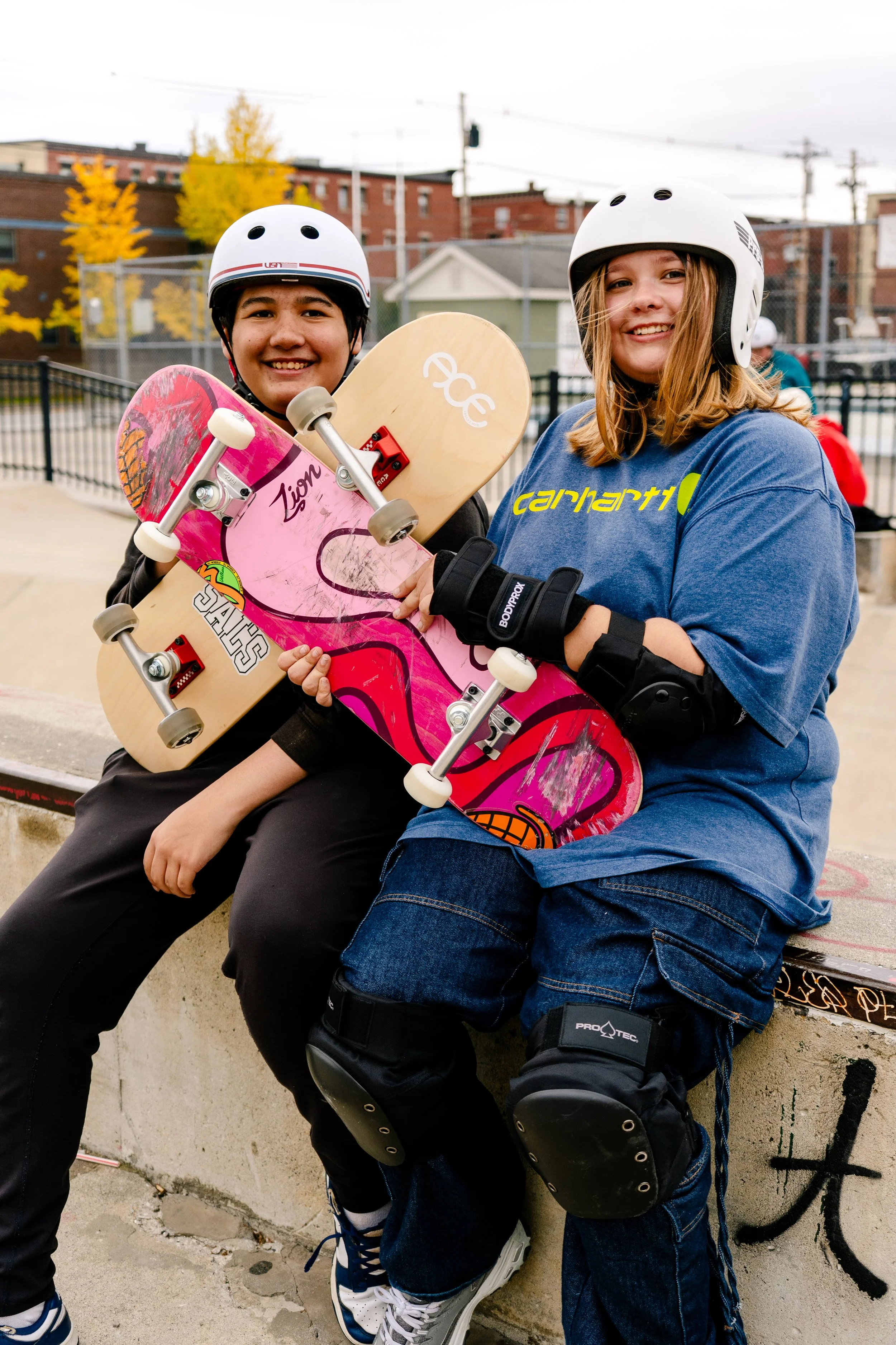 two skate to relate participants smiling in helmets and gear, sitting on a skate bench. they're holding up their boards to make an x.