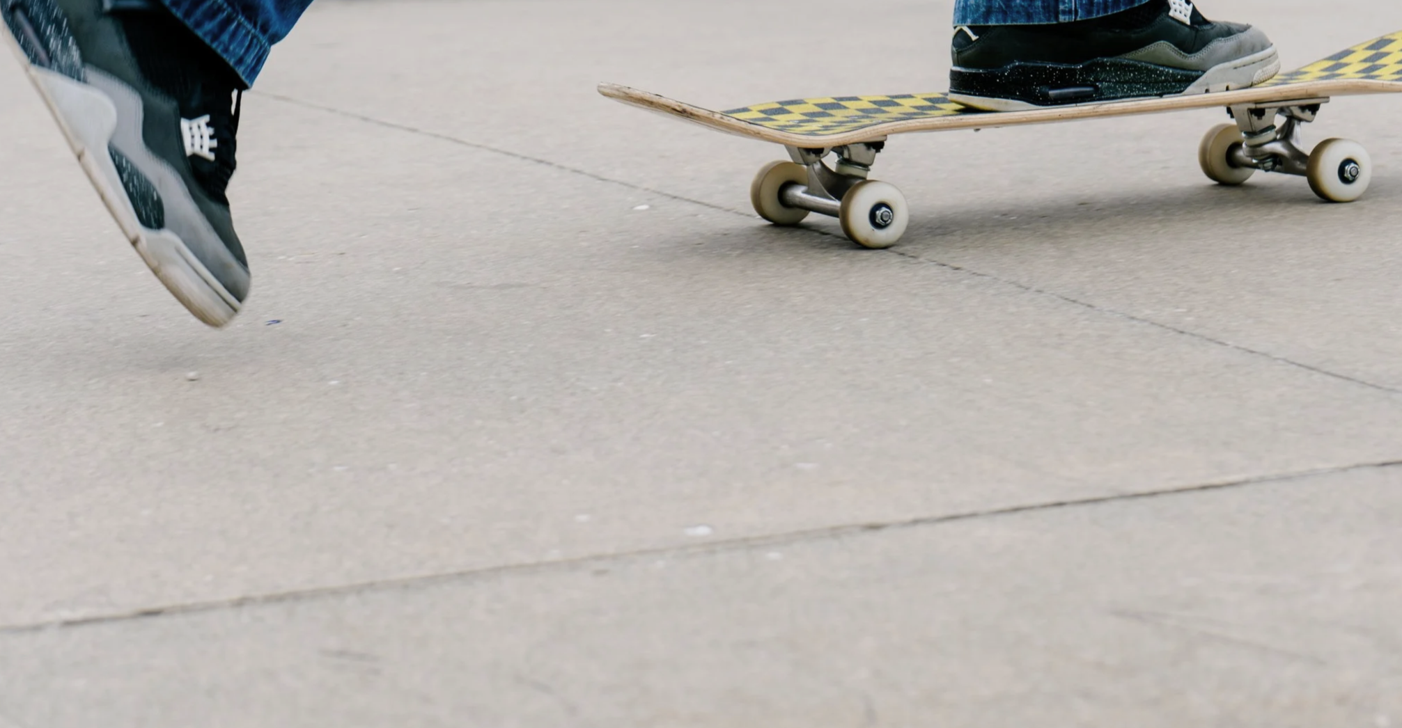 Close up image of someone pushing a yellow and black checkered skateboard on concrete. Only the feet and board are visible.