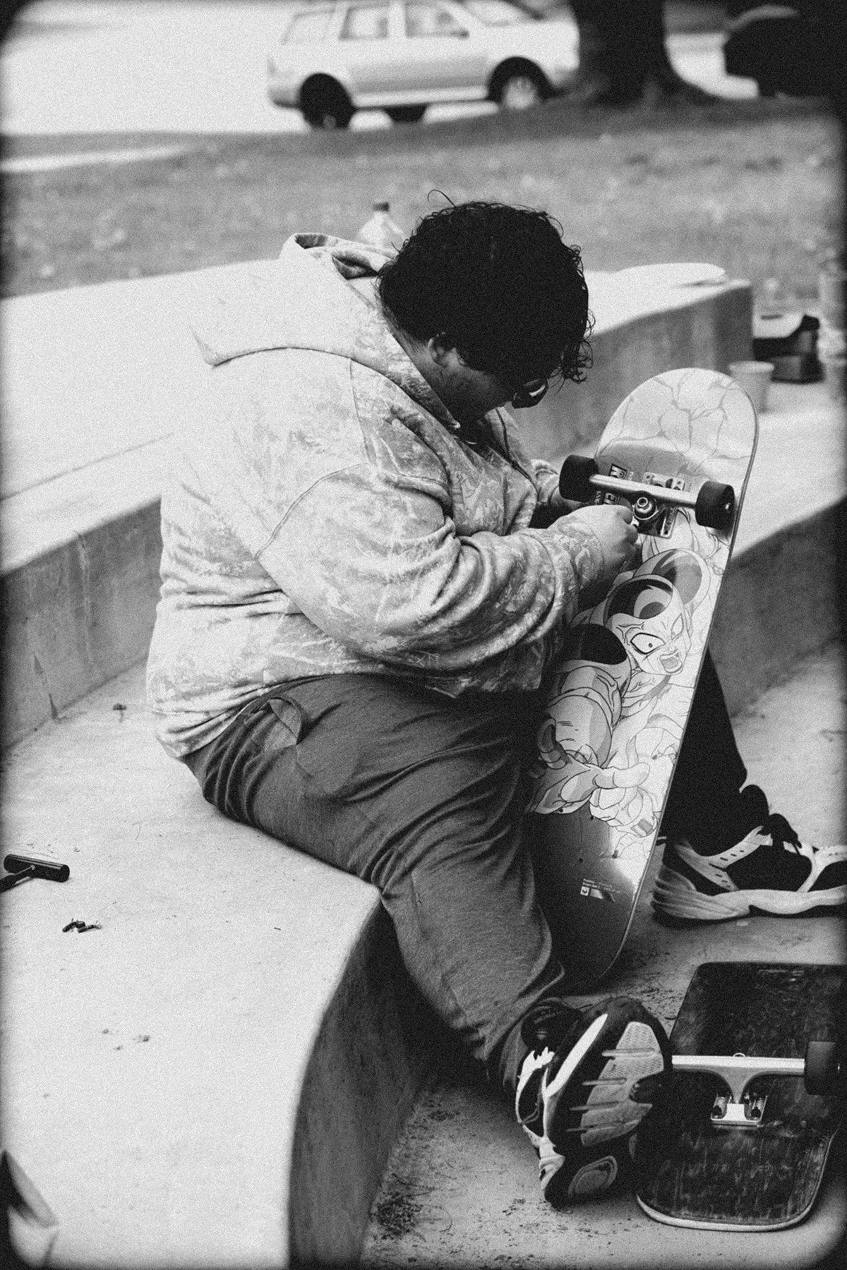 A young person with curly hair sitting on a skate park ledge, holding a skateboard with a cartoon character design.