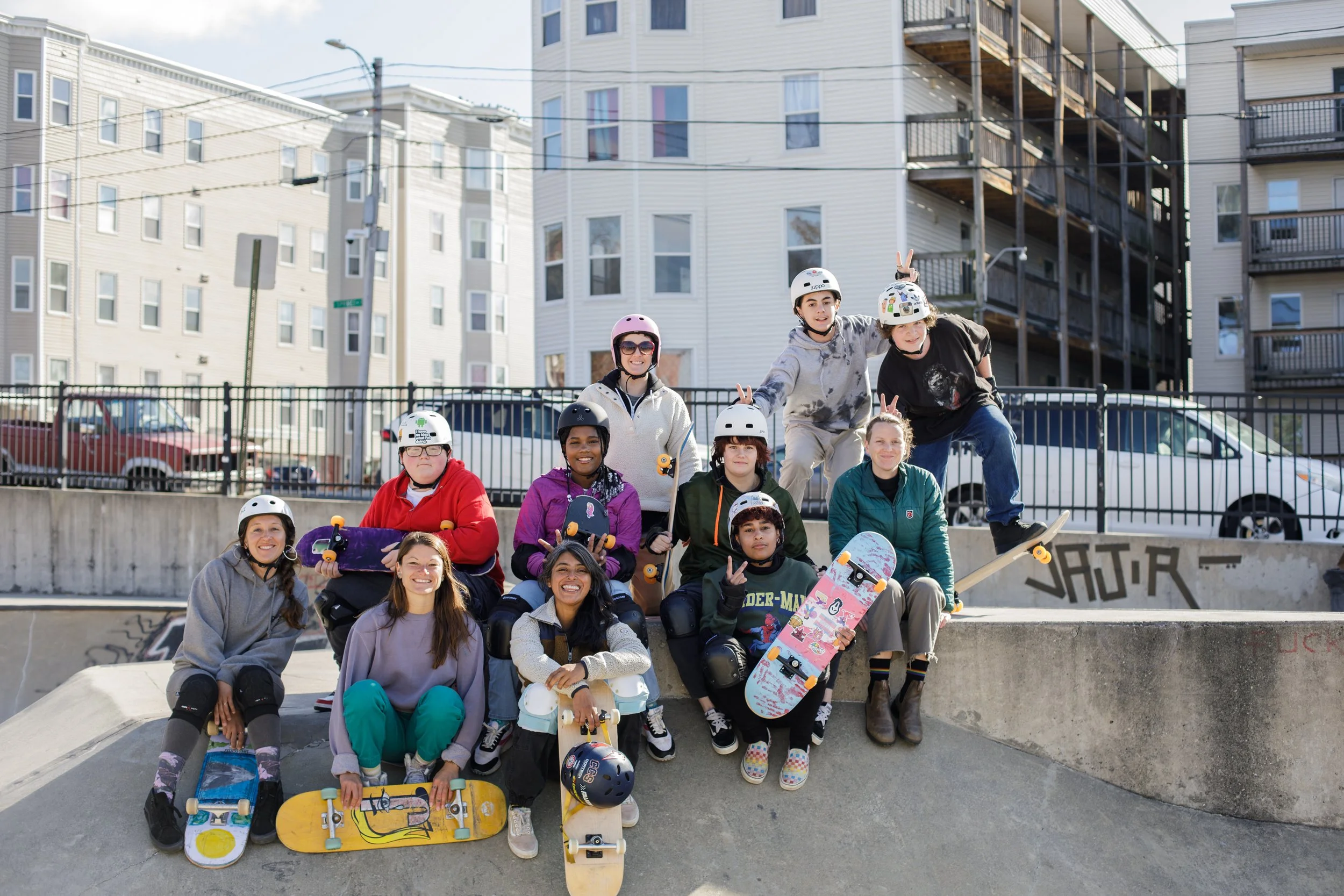 Group shot of skate to relate participants and mentors at a skatepark. Some are smiling, some are giving bunny ears, and most have on helmets.