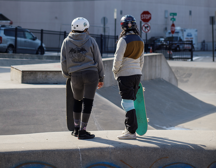 Two people in helmets with skateboards in hand standing, looking at a skatepark. Only their backs are visible.