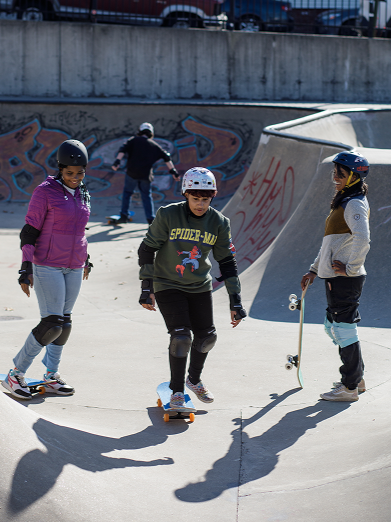 two youth members learning to skateboard at a skatepark, with a mentor on the side smiling.