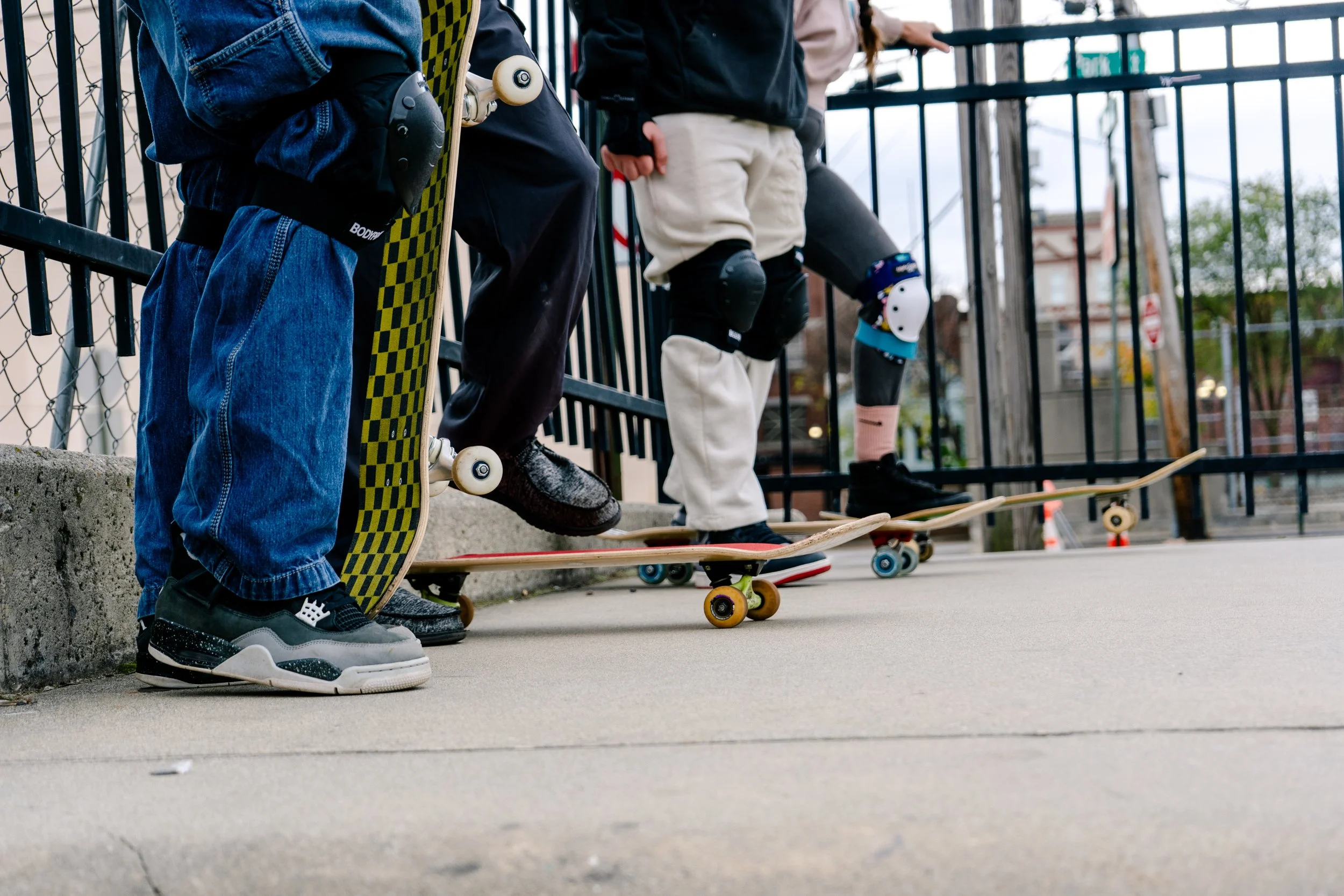 Several children, wearing protective kneepads, are standing on skateboards against a black metal fence in an outdoor urban setting.