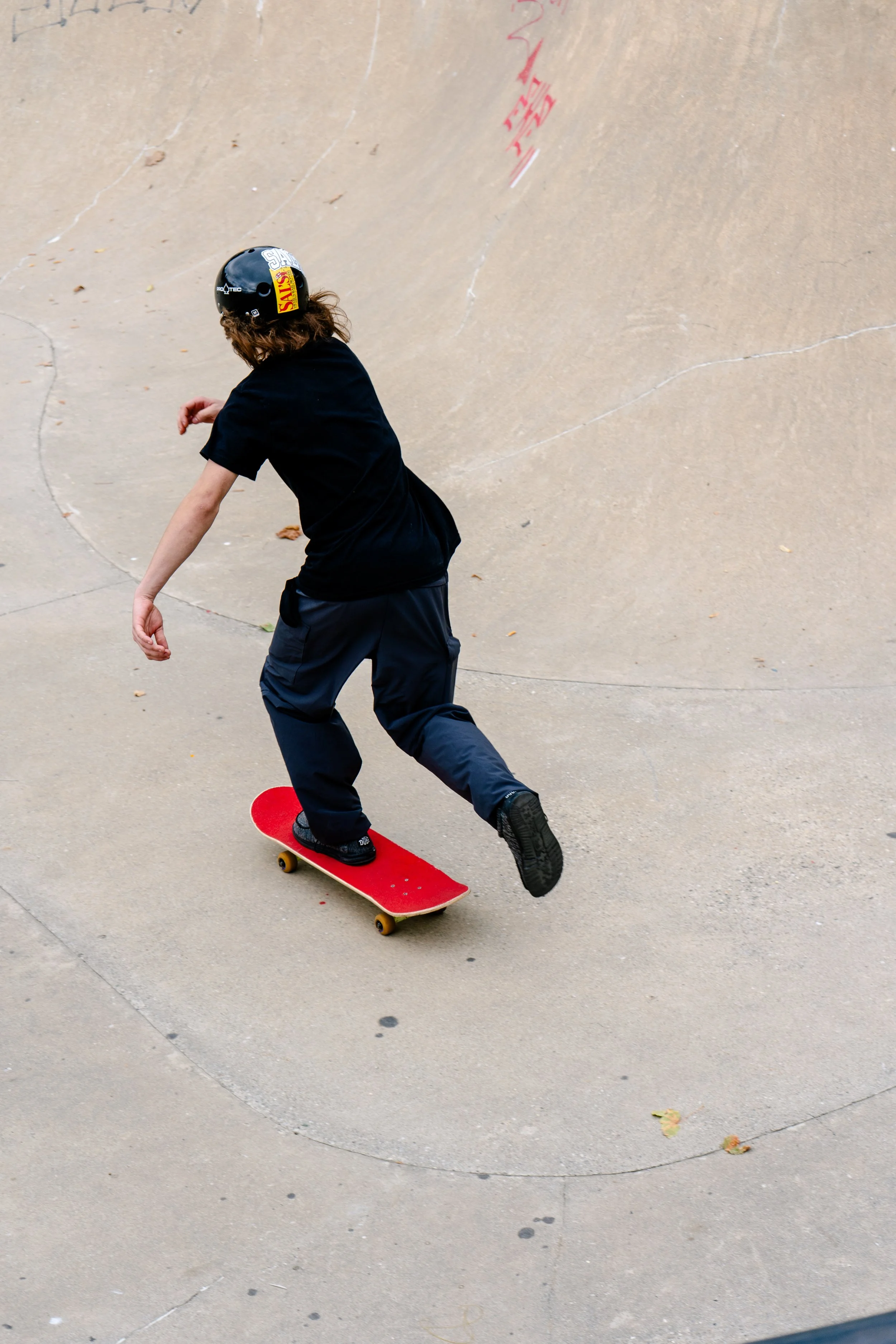 A person skateboarding at a skate park, wearing a black helmet, black t-shirt, and dark pants, with a brownish concrete background.