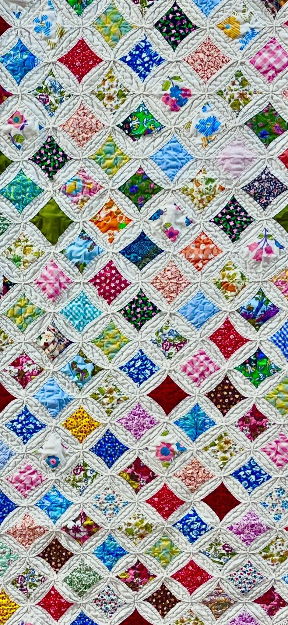A colorful patchwork quilt with a variety of floral and geometric fabric patterns arranged in a diamond grid, bordered with white quilting lines.