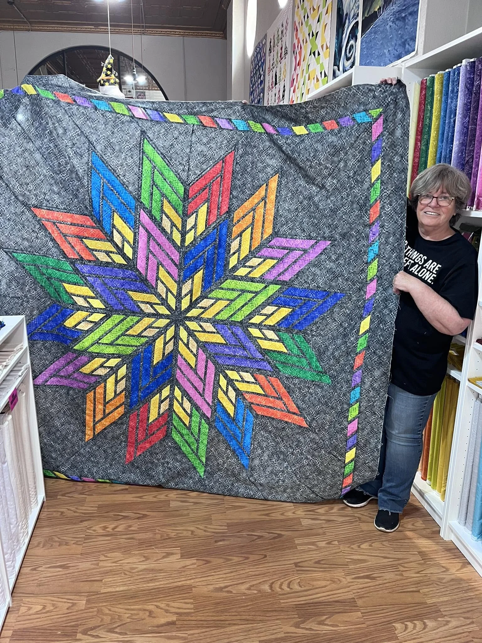 A woman with glasses, short gray hair, and a black t-shirt with white text, is holding a colorful quilt with a star design. The quilt has a gray background with a rainbow-colored star pattern and borders. She is standing in a fabric store with shelve
