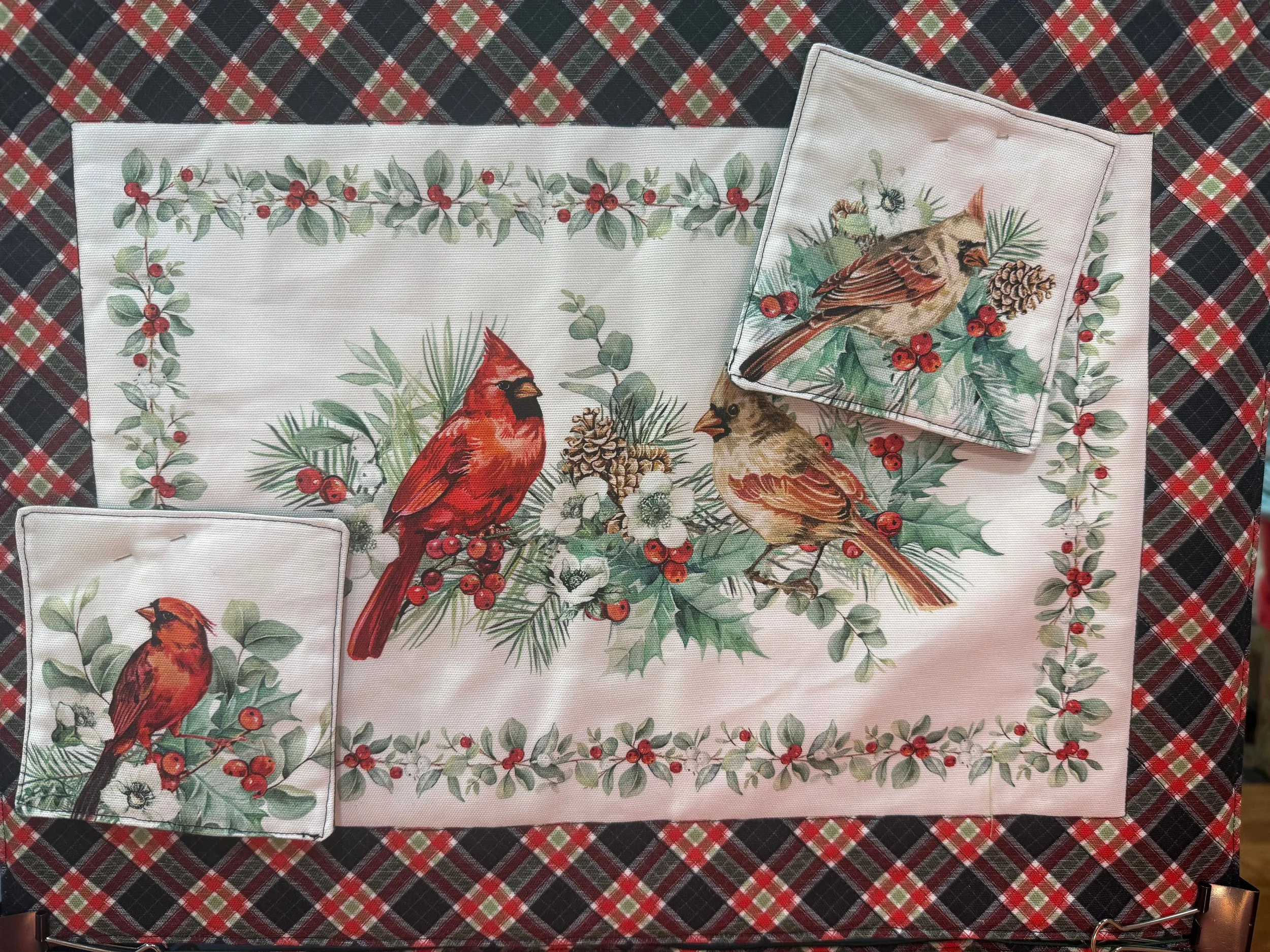 Christmas-themed table setting with a holiday placemat, napkins, and a decorative cloth featuring birds, pinecones, and holly berries.