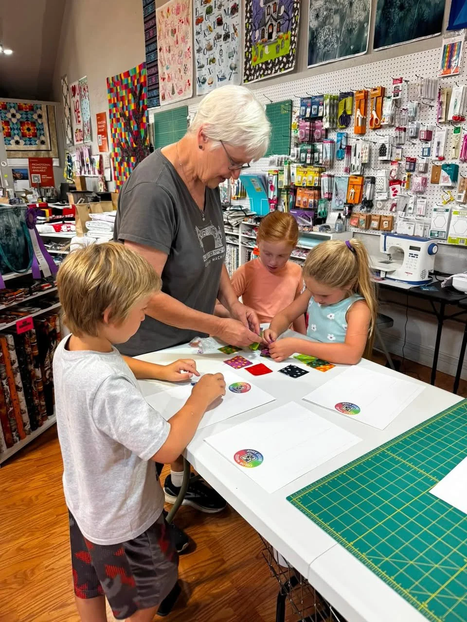 An elderly woman teaching three children how to quilt or sew at a craft store, with fabric and quilting supplies on the table and shelves of quilting materials in the background.