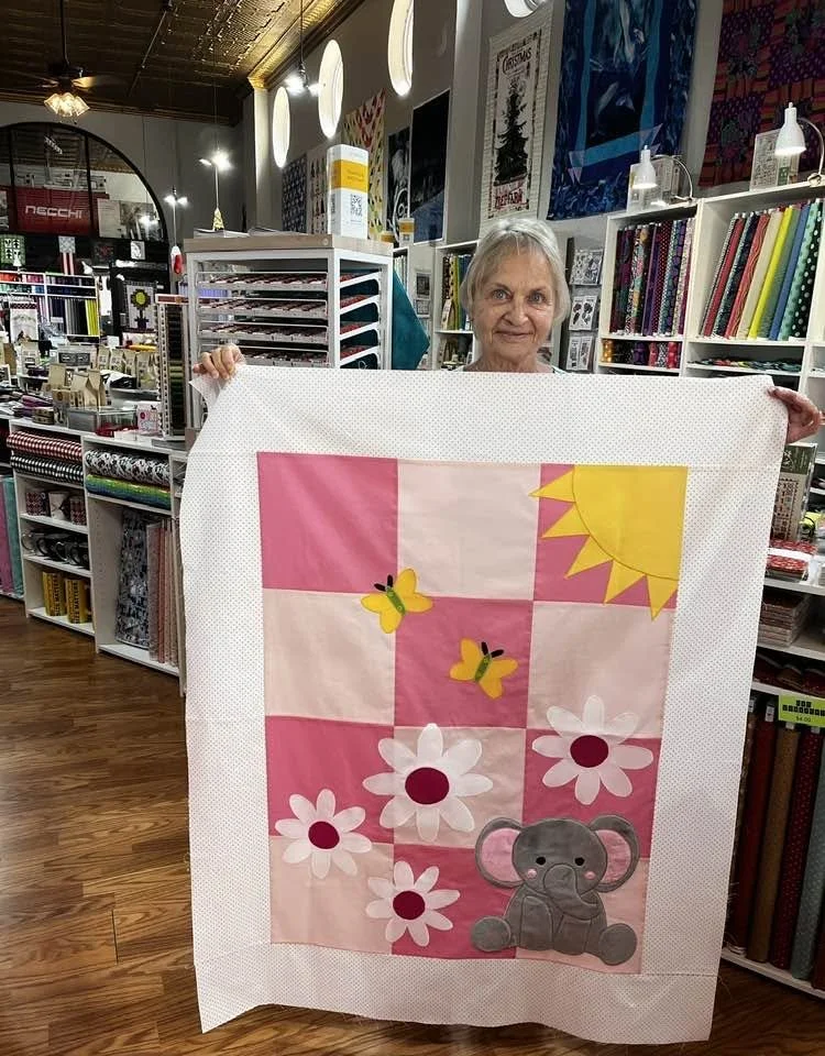 An elderly woman with gray hair holding up a colorful quilt in a fabric store. The quilt features a sun, butterflies, daisies, and an elephant on a pink and white patchwork background.