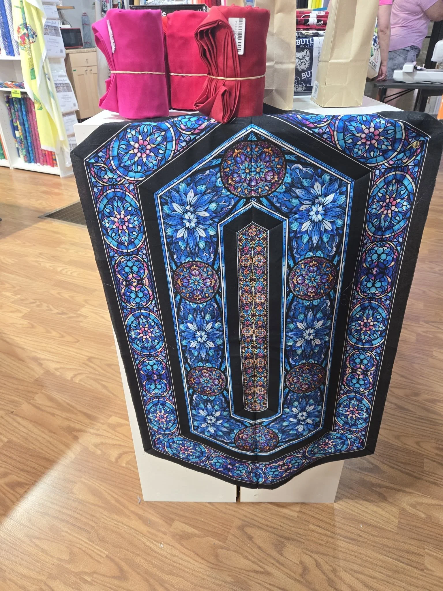 Display of brightly colored fabric items, including pink and red cloths, on a table with a decorative stained glass pattern tablecloth, inside a store with shelves of books or fabrics in the background.