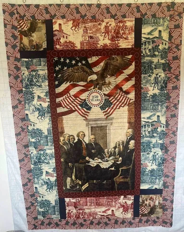 Patriotic quilt with a central image of the signing of the Declaration of Independence surrounded by images of American history, an eagle and American flags, celebrating 250 years.