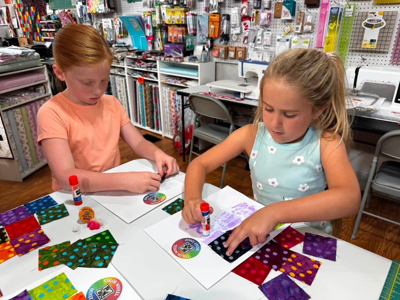 Two young girls are sitting at a craft table in a store, working on fabric projects with glue sticks. The table is covered with colorful fabric squares and crafting supplies, and the background features shelves of craft materials and sewing equipment