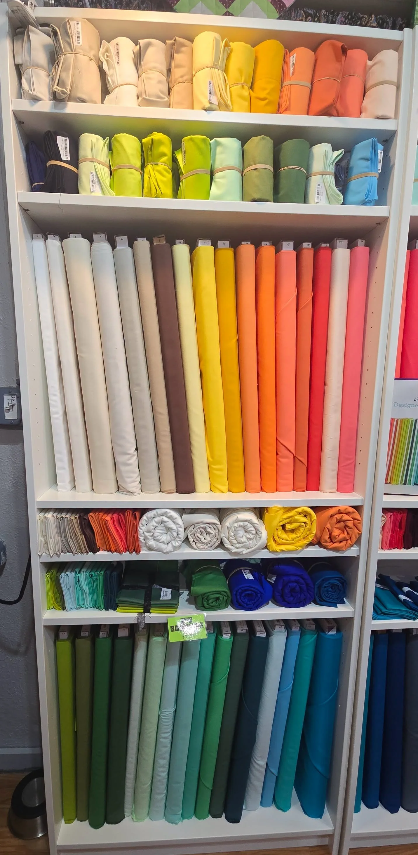 Shelving unit holding various colorful fabric rolls organized by color in shades of beige, orange, yellow, green, blue, and teal.