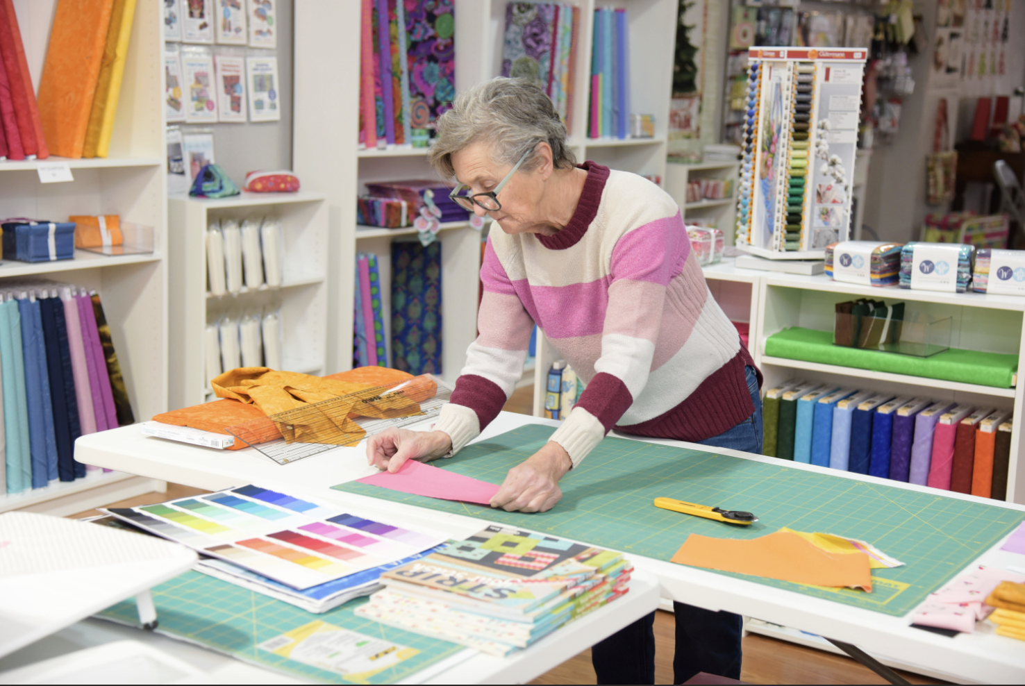 A woman working at a sewing or craft table in a fabric store, surrounded by fabric samples, color swatches, and pattern books.