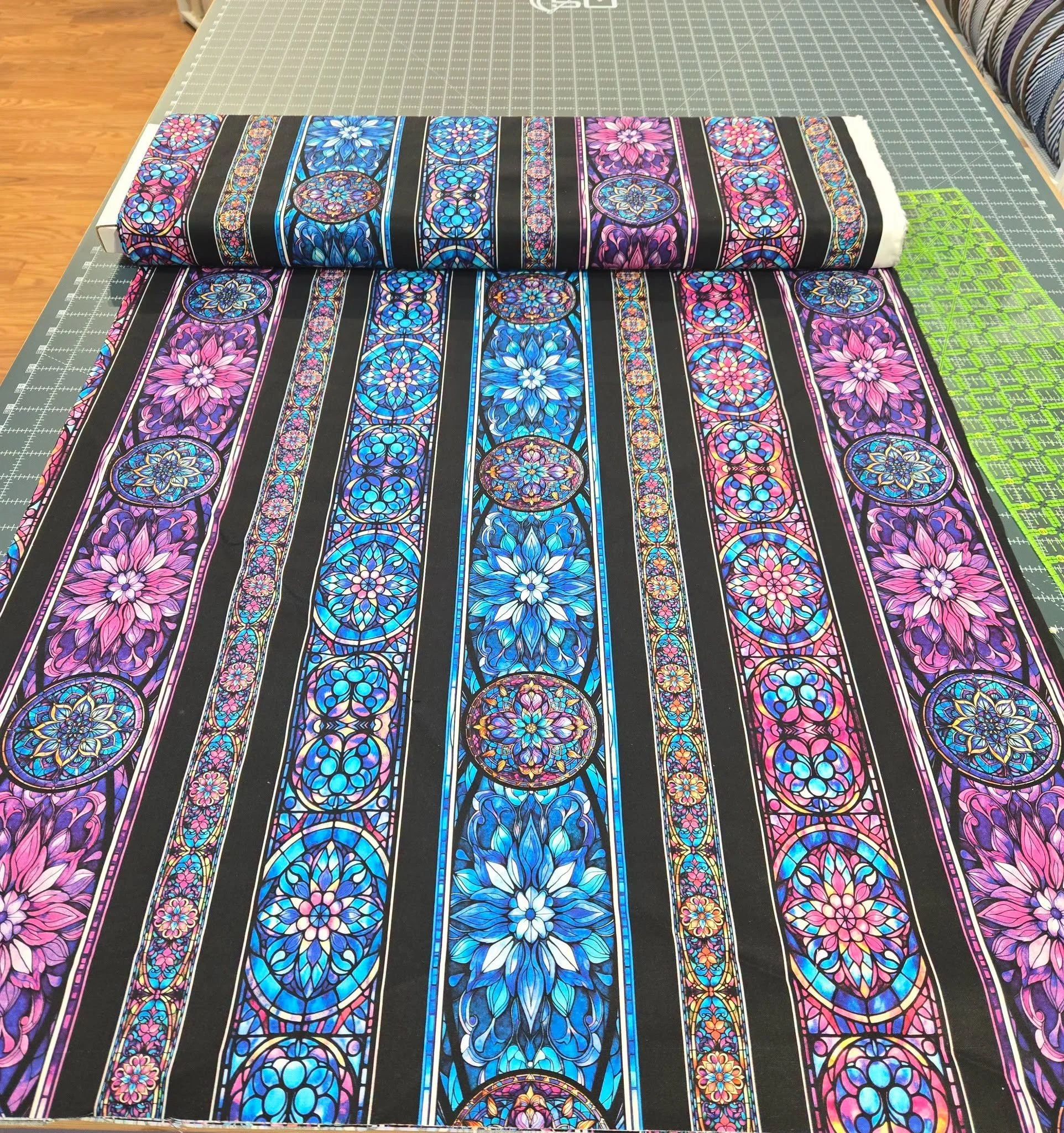 A roll of fabric with a black background and colorful stained glass-like floral patterns in pink, blue, purple, and white, placed on a green cutting mat on a worktable.