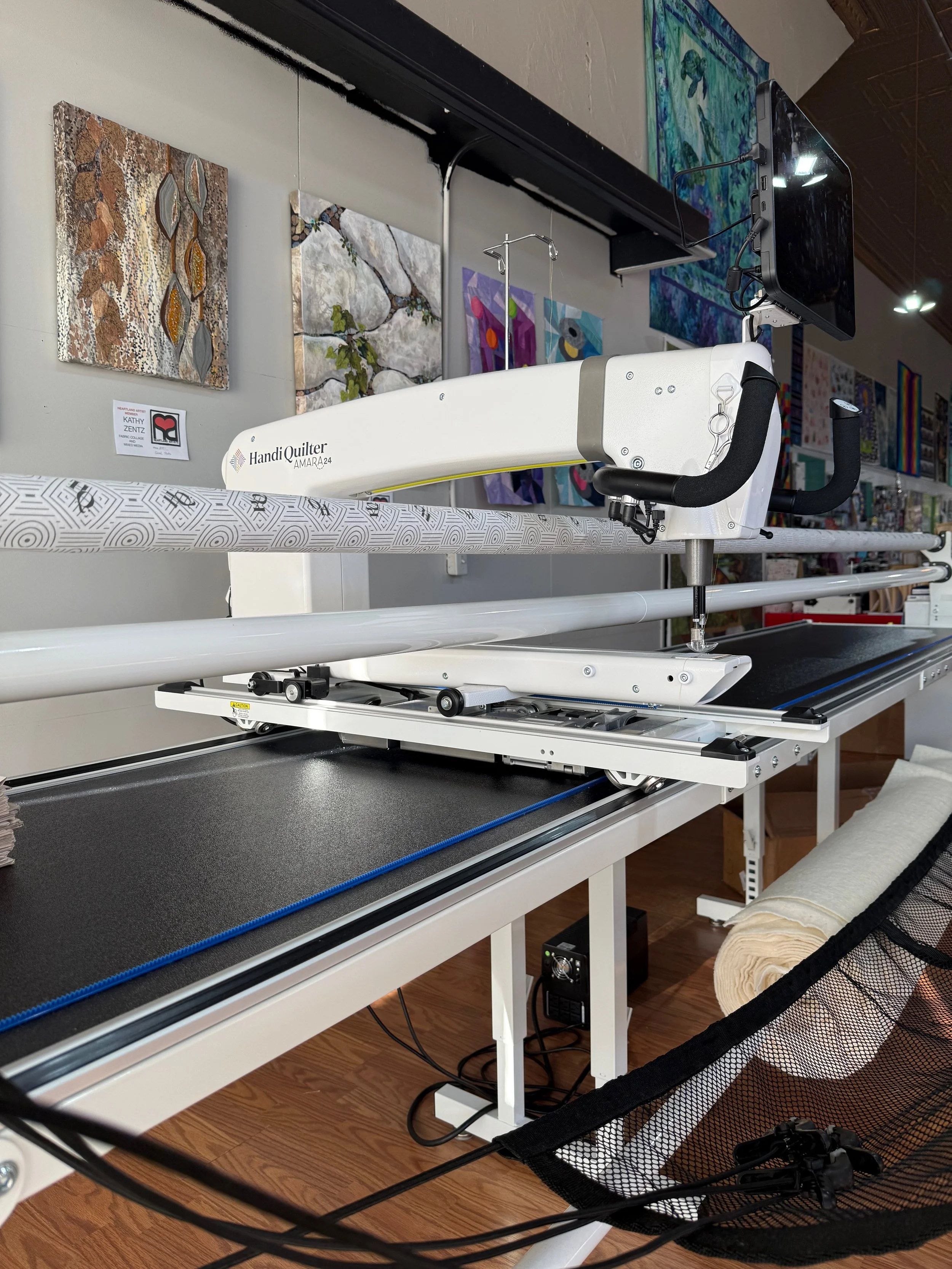 A quilting machine with a computer screen, surrounded by colorful fabric art and a wooden floor.