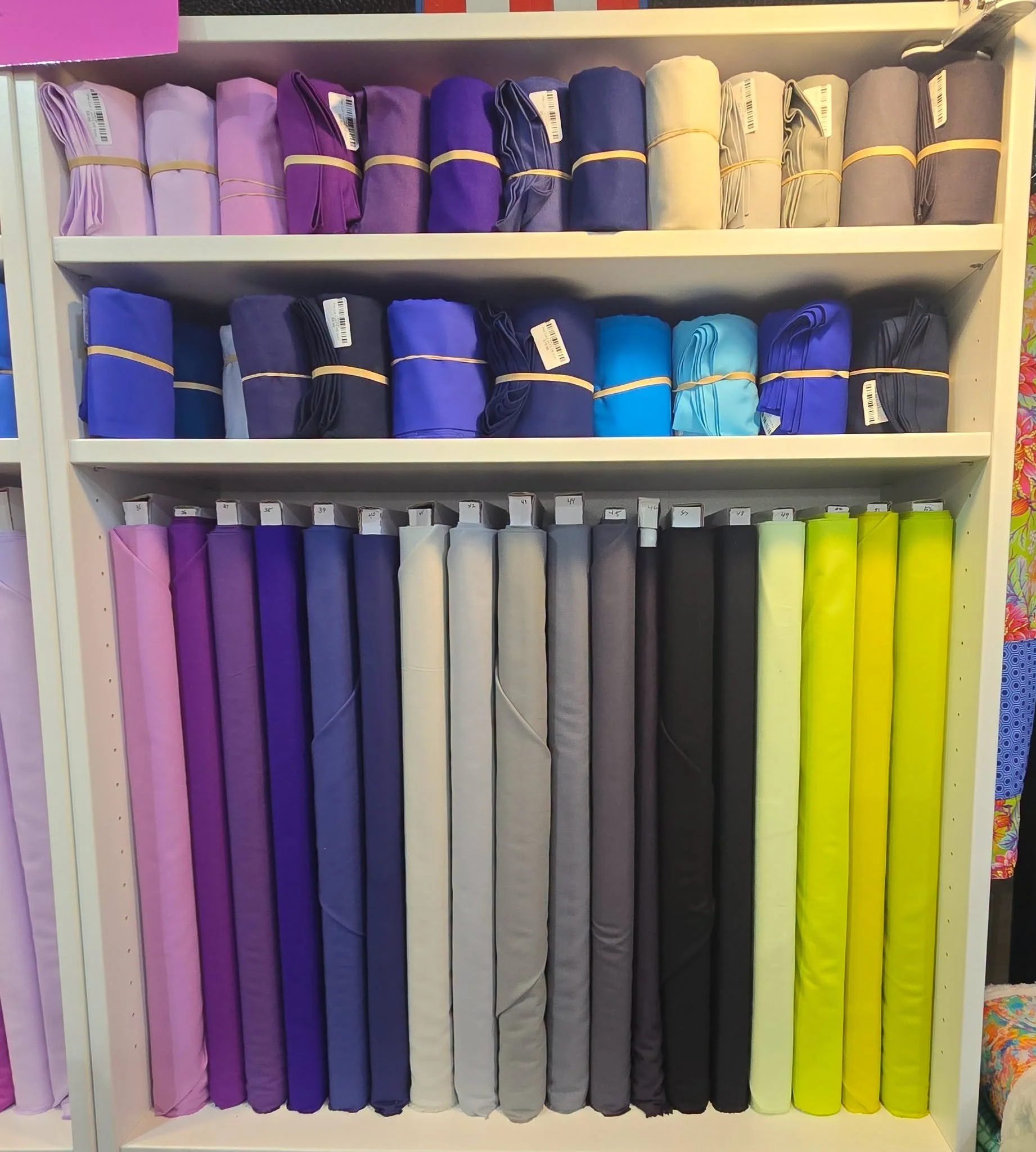 Shelf filled with rolled-up fabrics or textiles in shades of pink, purple, blue, gray, and yellow.
