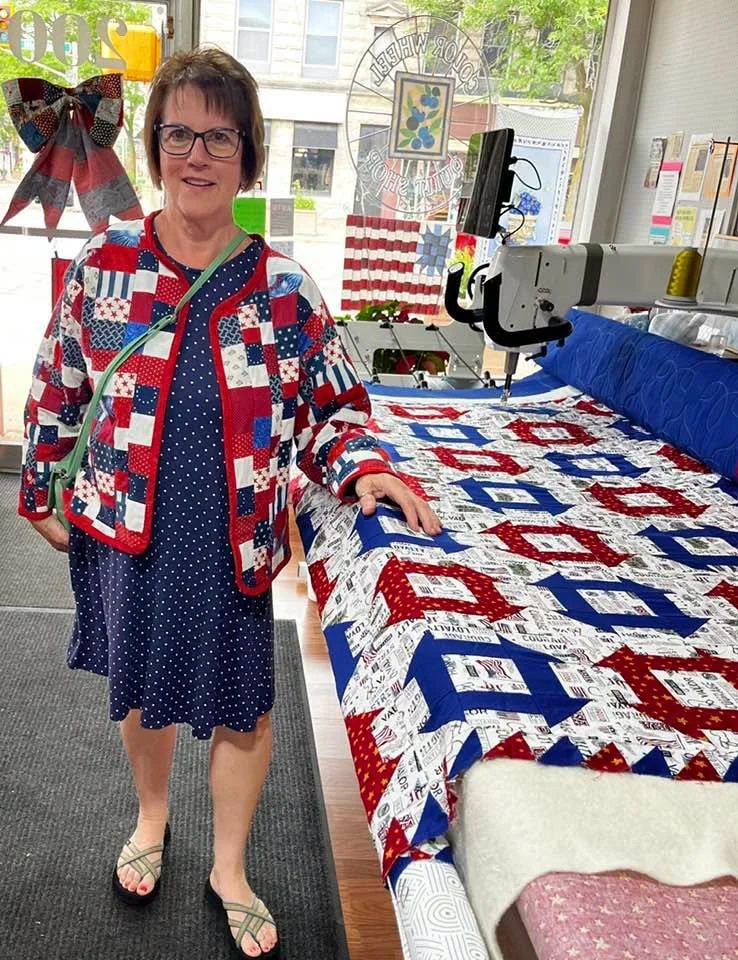 Woman in patriotic-themed clothing standing in quilting shop with sewing machine and quilt.