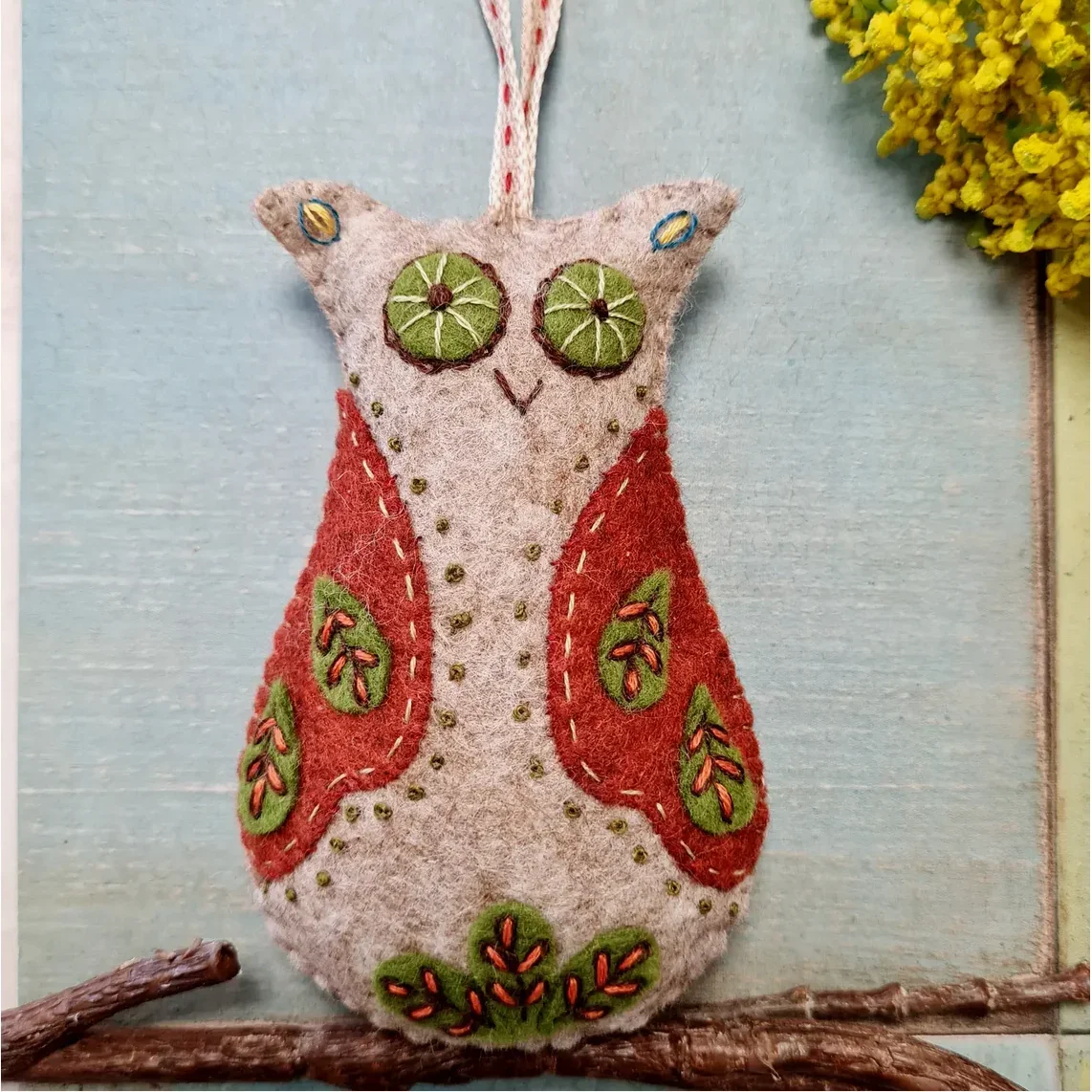 Felt owl ornament with embroidered details and seed eye accents, hanging on a wall with a branch at its base and a yellow floral arrangement nearby.