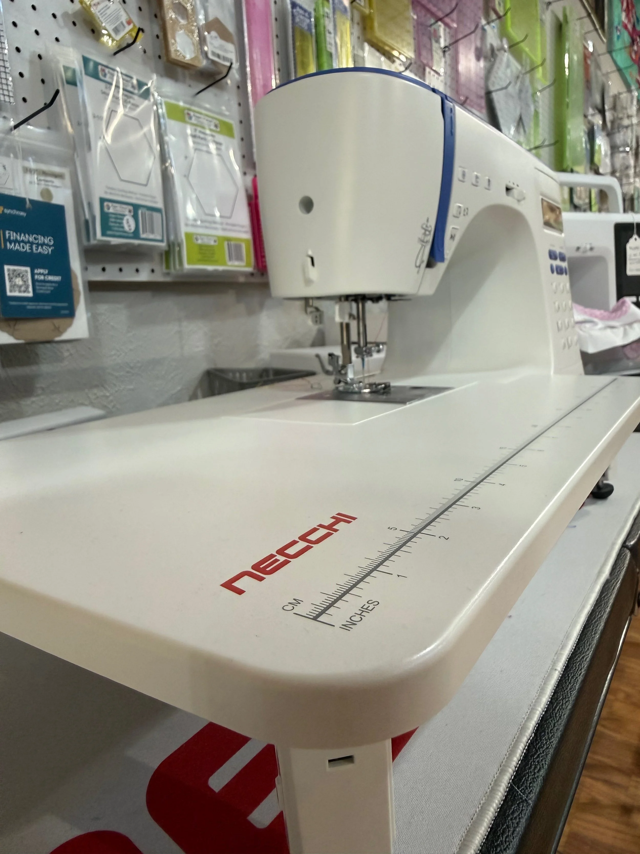A Necchi white sewing machine with a built-in ruler in centimeters and inches, placed on a surface in a store, with various sewing accessories and supplies on shelves in the background.