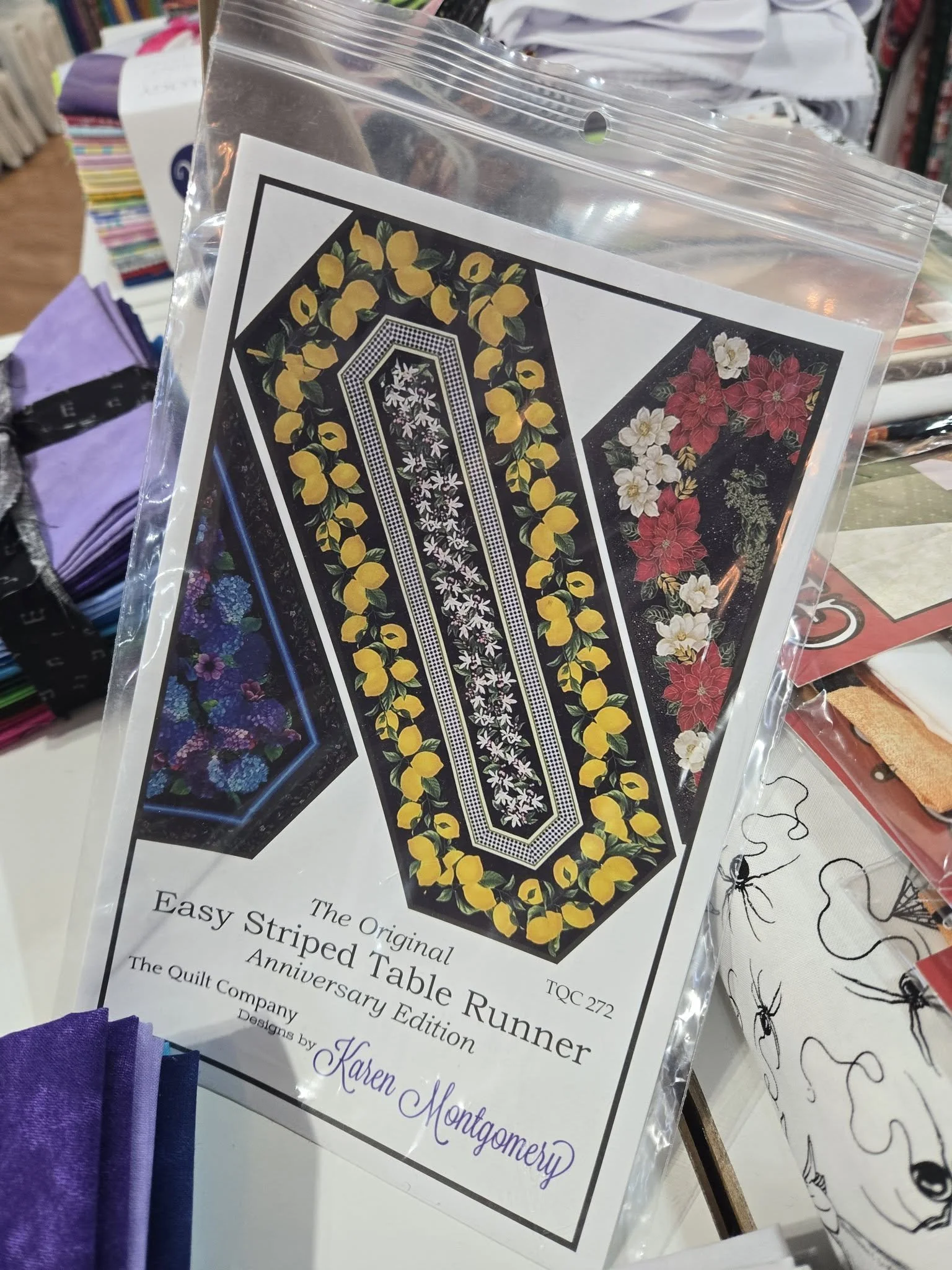 A clear plastic bag contains a printed fabric pattern featuring yellow lemons and green leaves on a black background, with an inset pattern of white floral design on a black background, for quilting or sewing projects.