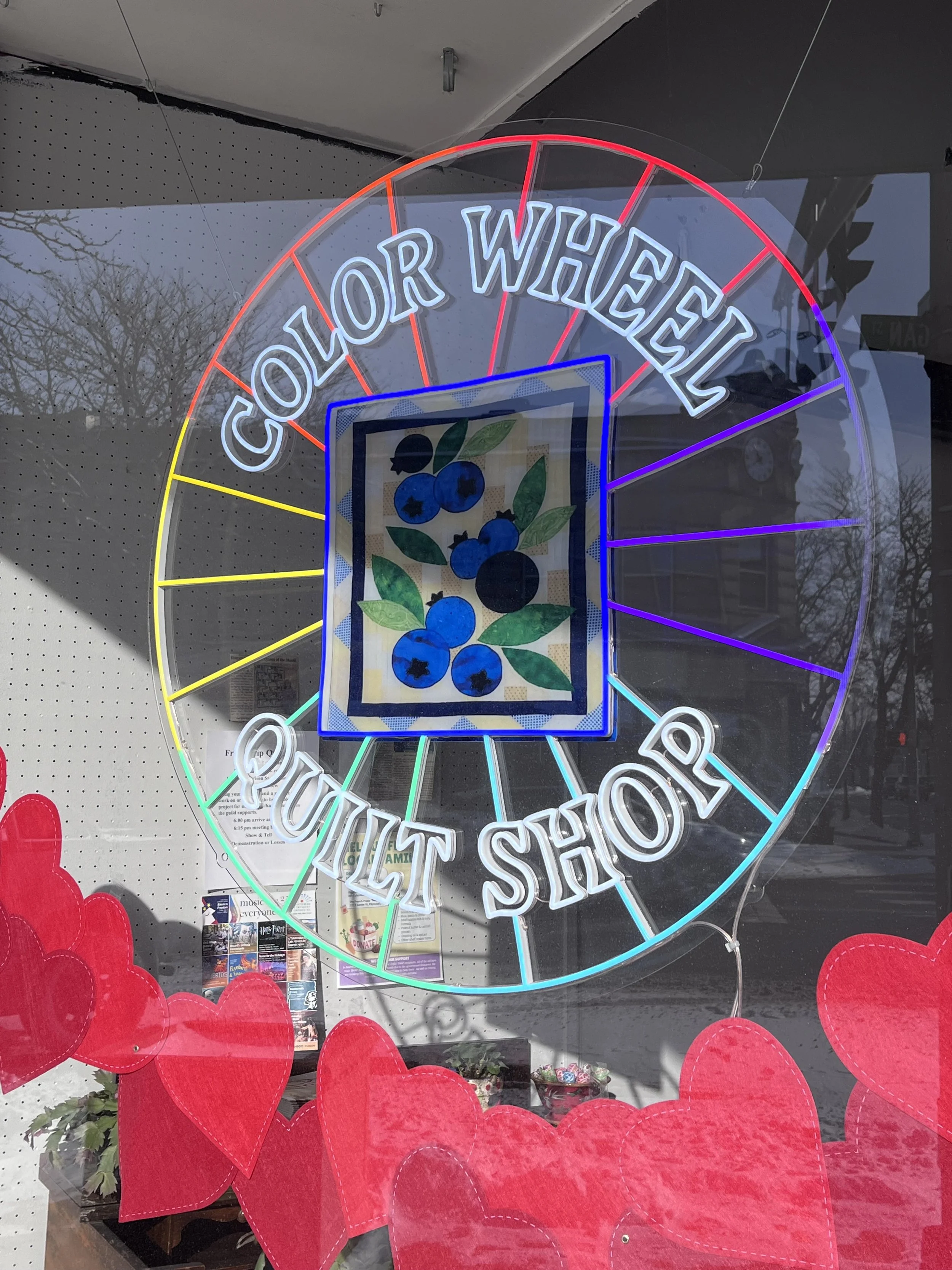 Store window display with a neon sign that reads 'Color Wheel Shop' and a display of red paper hearts hanging in front.