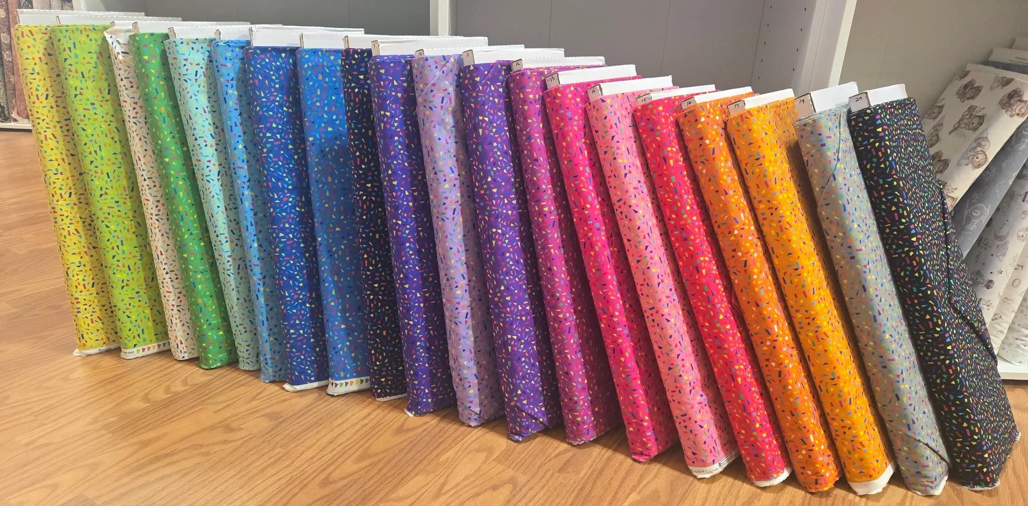 A display of colorful wrapping paper rolls arranged in a row on a wooden surface, featuring vibrant hues and confetti-like patterns.