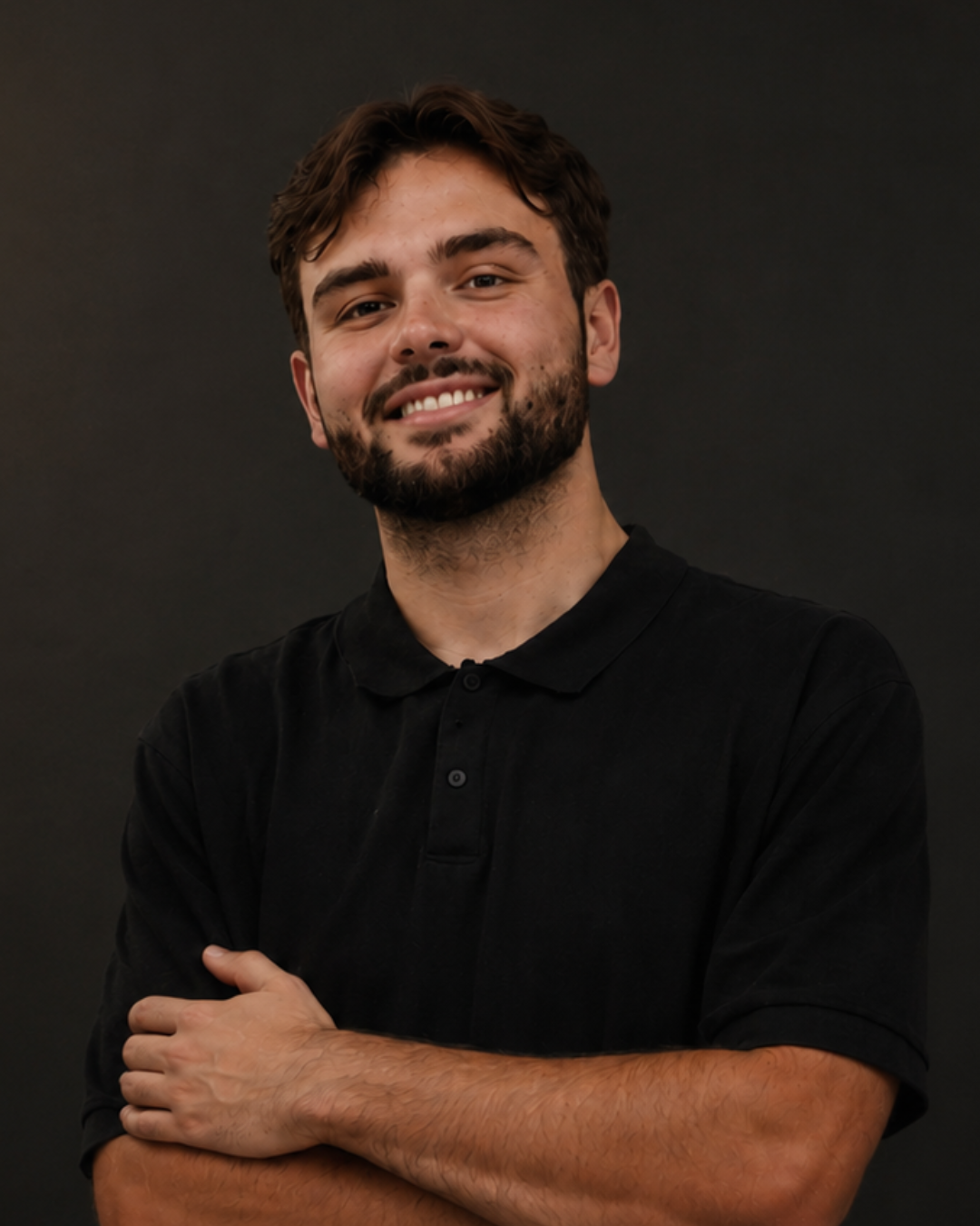 A young man with dark hair, a beard, smiling, wearing a black shirt, standing against a plain dark background with arms crossed.