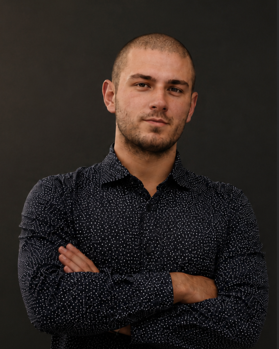 A young man with short hair and a beard, wearing a black shirt with white polka dots, standing against a plain dark background with his arms crossed.