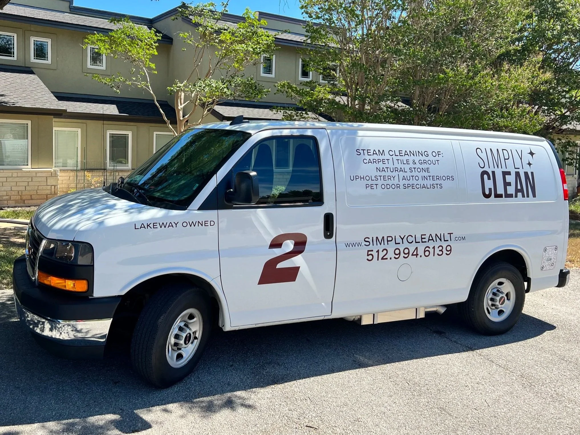 White van parked on a residential street with trees and multi-story houses in the background. The van has signage indicating cleaning services, including steam cleaning, upholstery, pet odor removal, and other cleaning services.