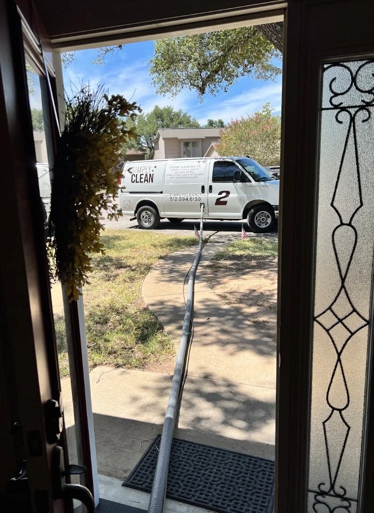 A view from inside a house looking out the front door onto a sidewalk and lawn. Outside, a cleaning service van with the logo 'Simply Clean' and the words 'Steam Cleaning' is parked on the street. A long hose extends from the house to the van, likely connected to cleaning equipment inside.