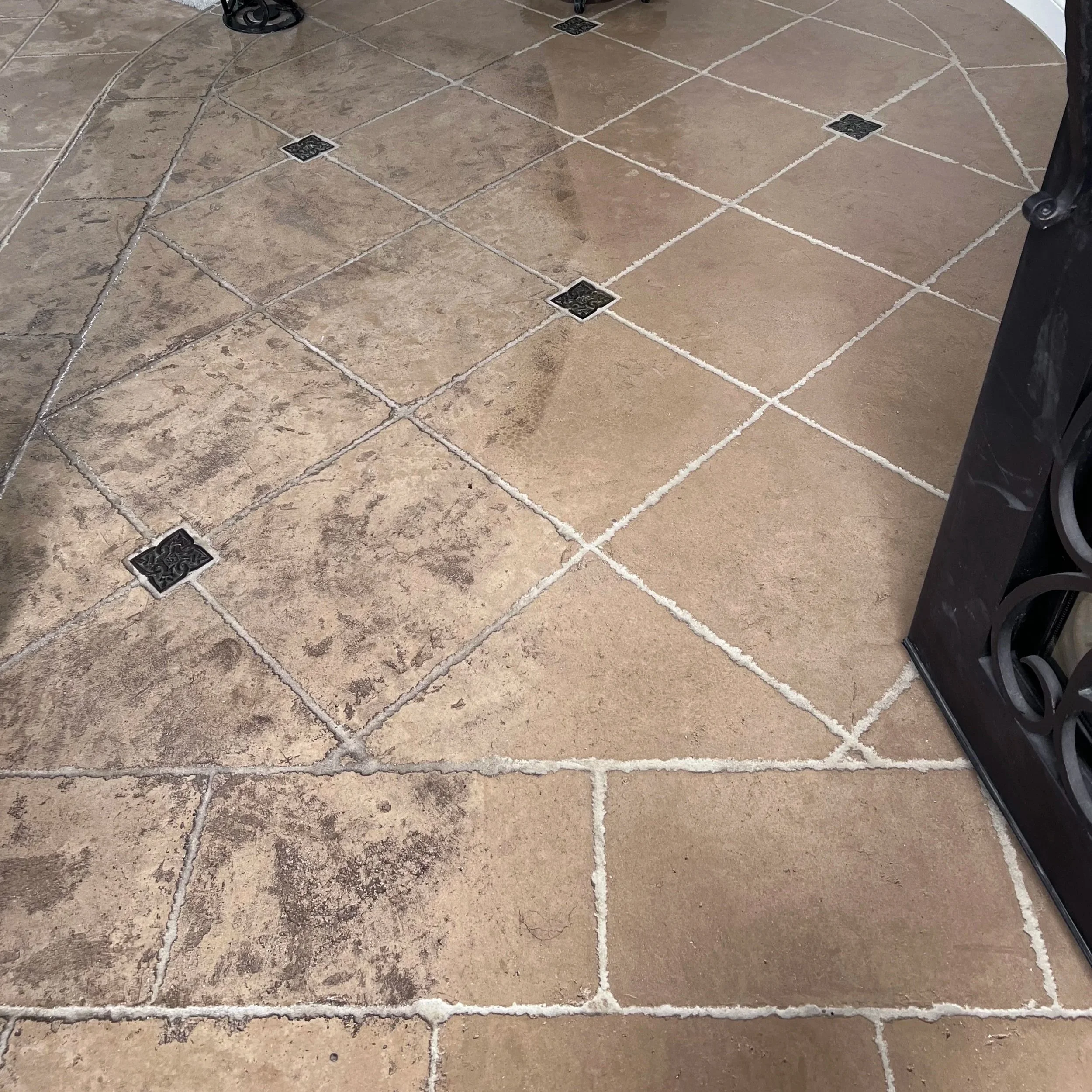 Close-up view of a tiled floor with large beige tiles, black decorative floor drains, and dark-colored furniture or appliance part at the edge.