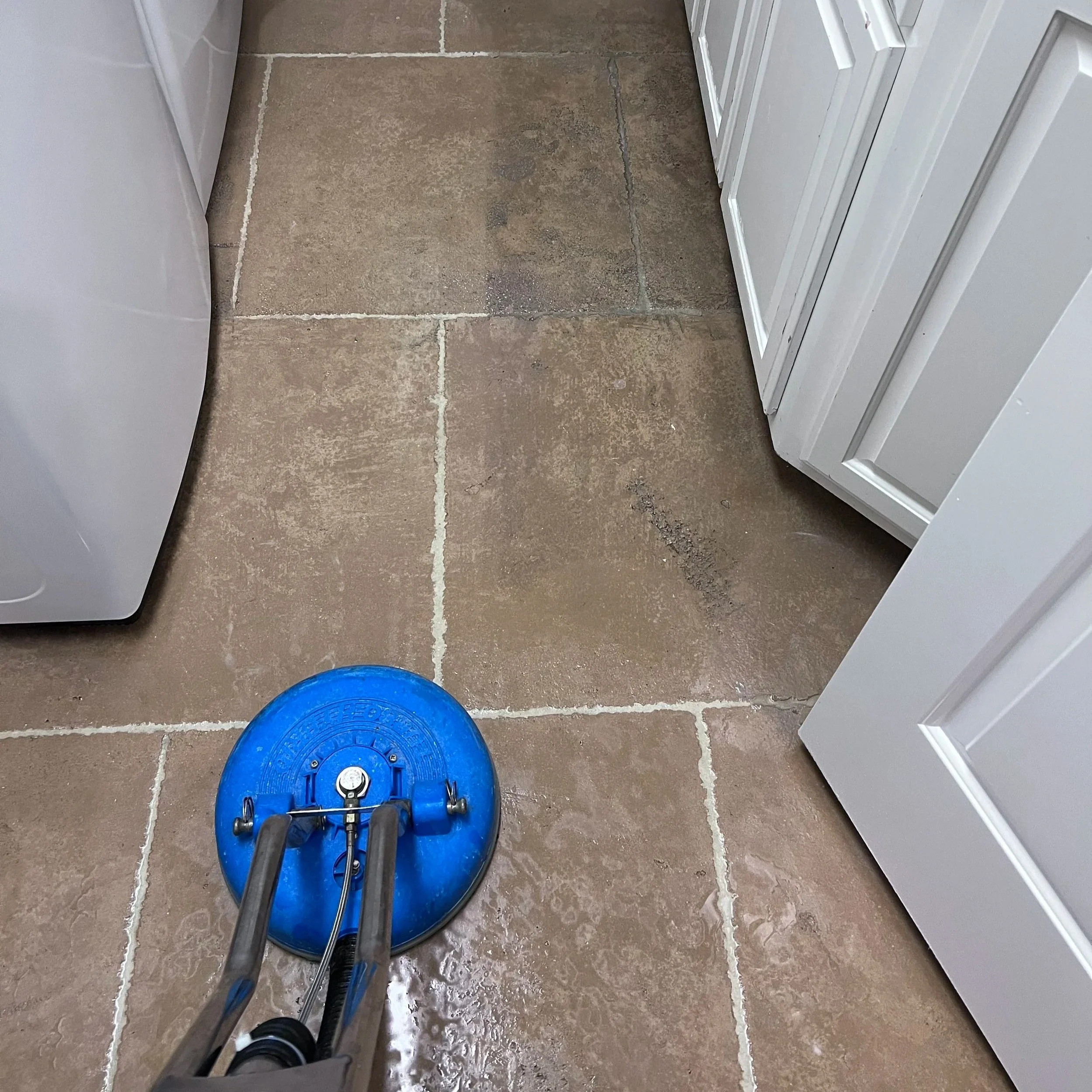 Person using a blue spinner to wash the floor with water splashing. The floor is tile and appears to be in a kitchen or laundry room, with white cabinets on the right side.