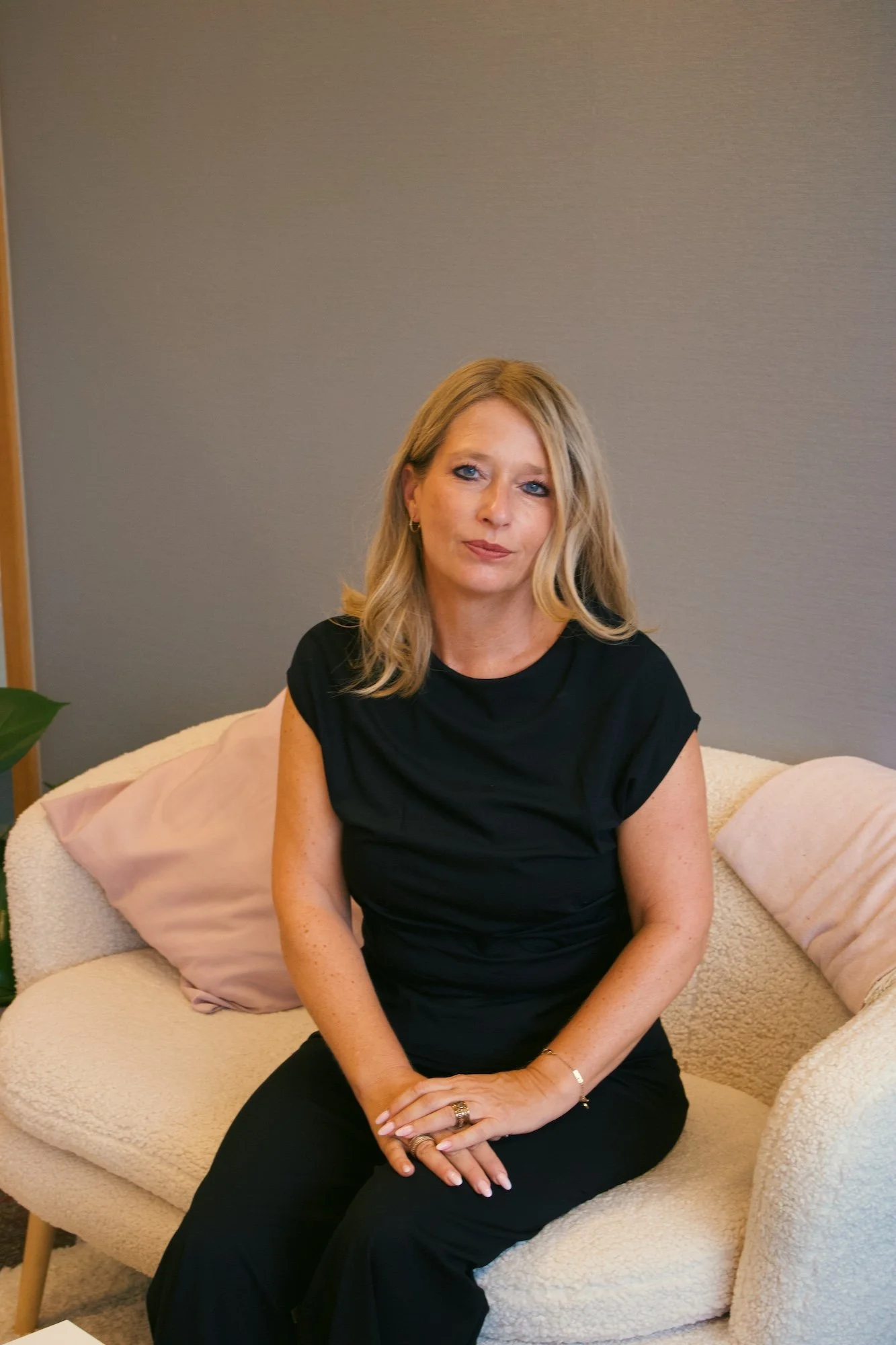 A woman with blonde hair sitting on a beige couch with pink pillows, wearing a black top and black pants, with her hands resting on her lap, in a room with a plain gray wall.