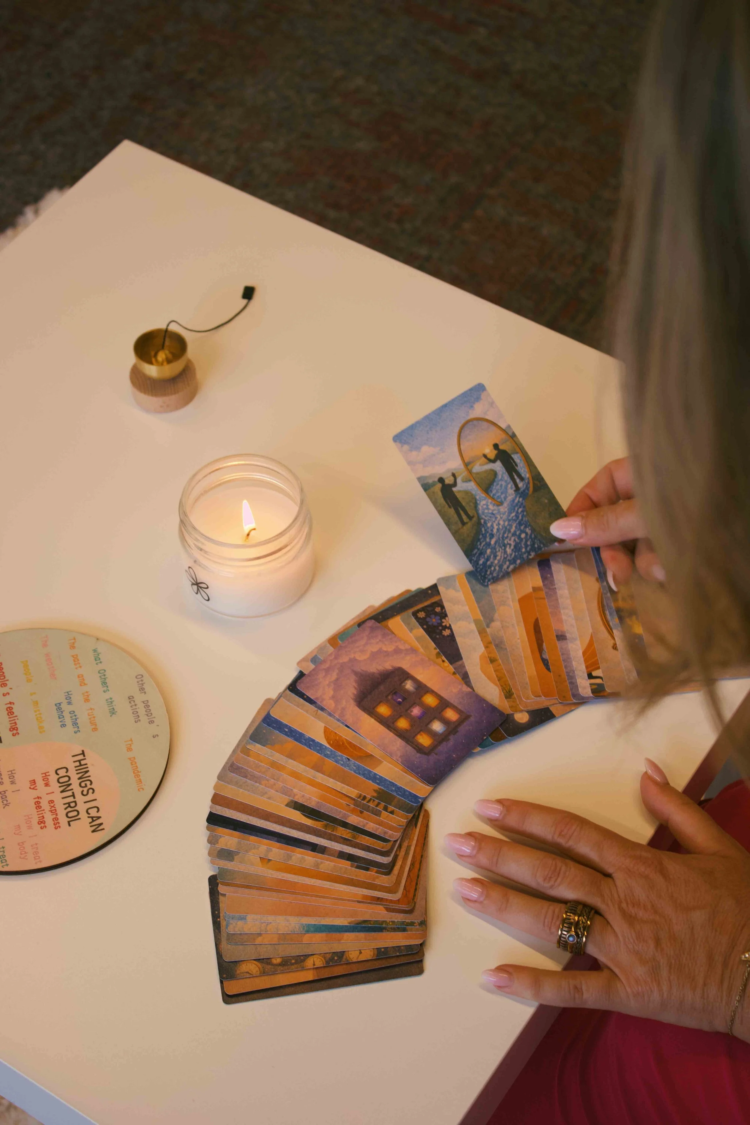 A person holding a deck of tarot cards over a table with a lit candle, a bowl, and a circular card with text, suggesting a tarot reading or spiritual practice.