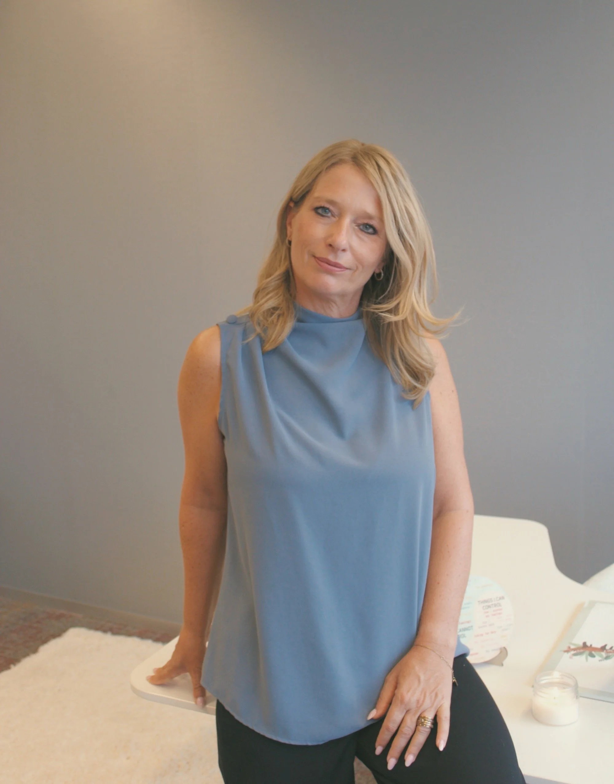 A woman with blonde hair wearing a light blue sleeveless top sitting at a table with a candle and informational signs.