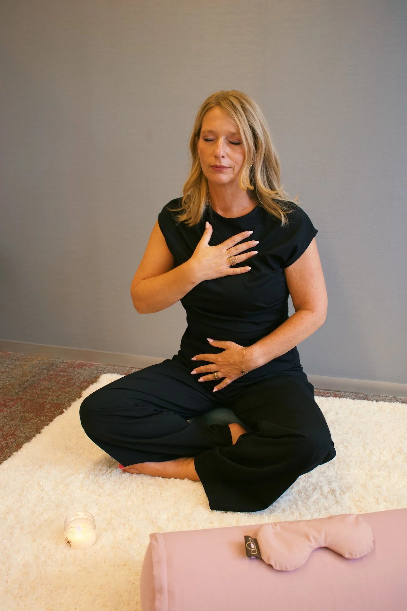 A woman in a black outfit sits cross-legged on a white fluffy rug, with one hand over her chest and the other on her stomach, eyes closed, in a meditative pose, in a room with gray walls, a lit candle, and a pink massage table with a pink cloth and a pink eye pillow nearby.