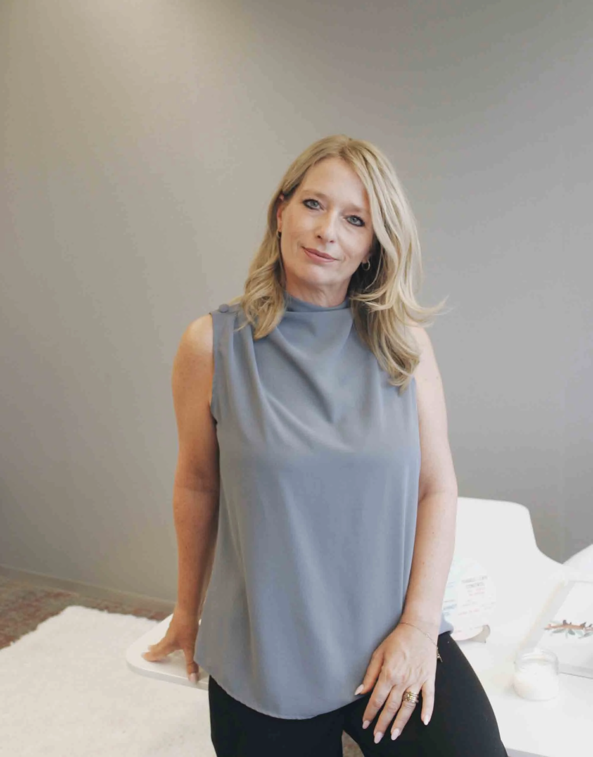 A woman with blonde hair wearing a gray sleeveless top, standing indoors near a white table, with a neutral gray wall in the background.