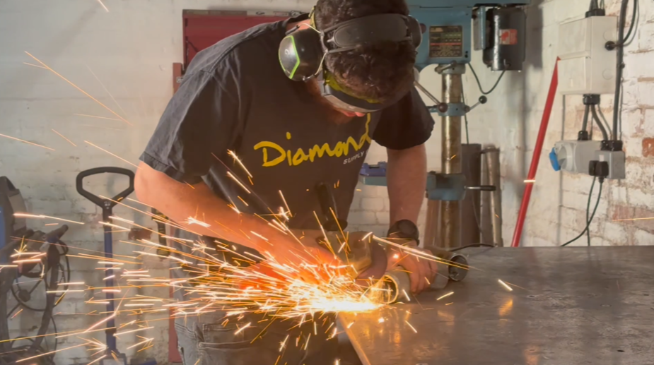 A person wearing safety goggles and headphones using a power tool to cut metal, sparks flying in a workshop.