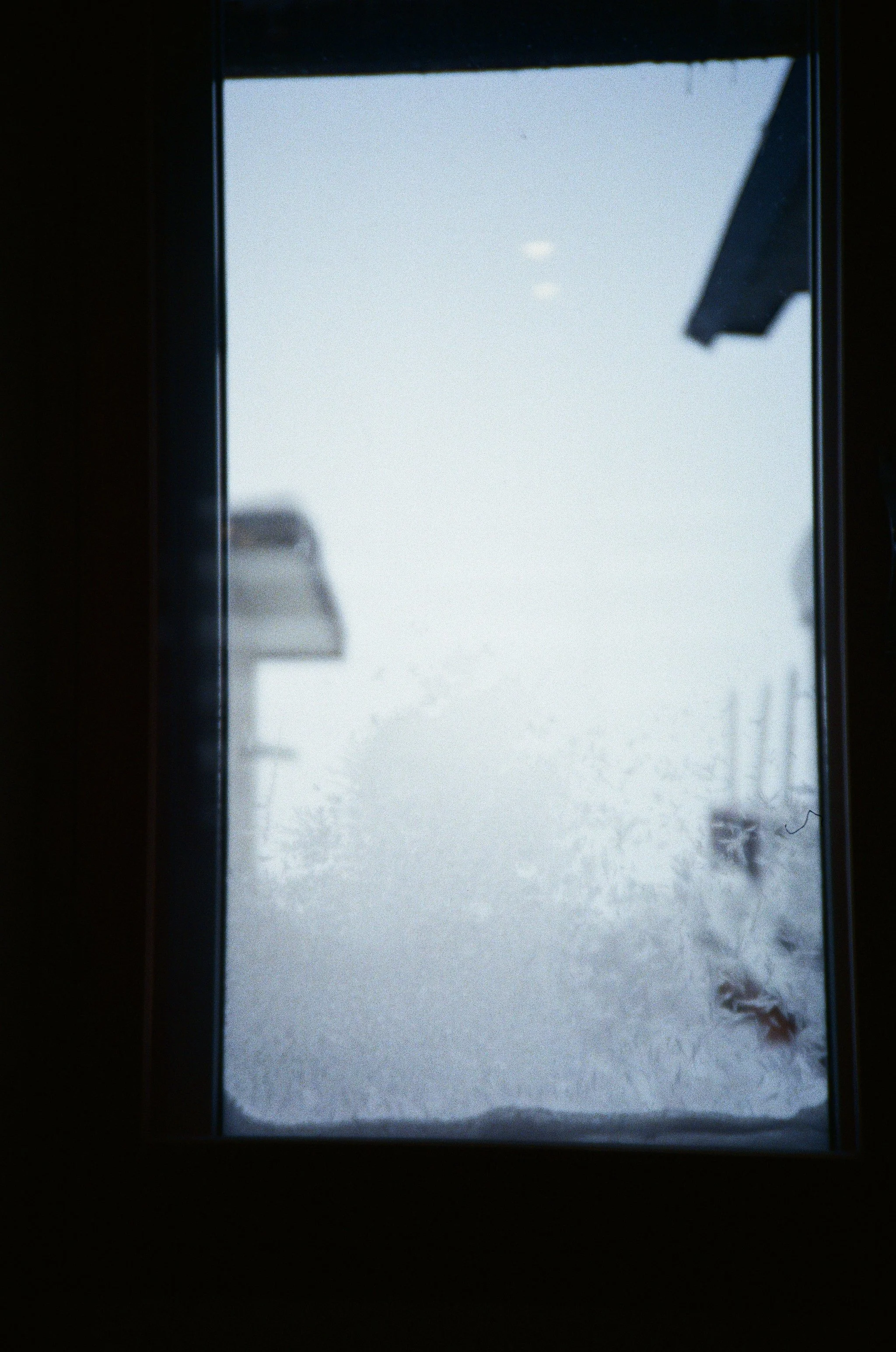 Looking through a window at a snowy landscape with a snow-covered road, houses, and a ski lift in the distance, under a cloudy sky.