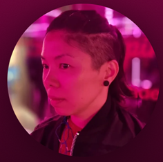 A person with a shaved side hairstyle in a dark jacket, wearing earrings, in front of pink and purple lighting.