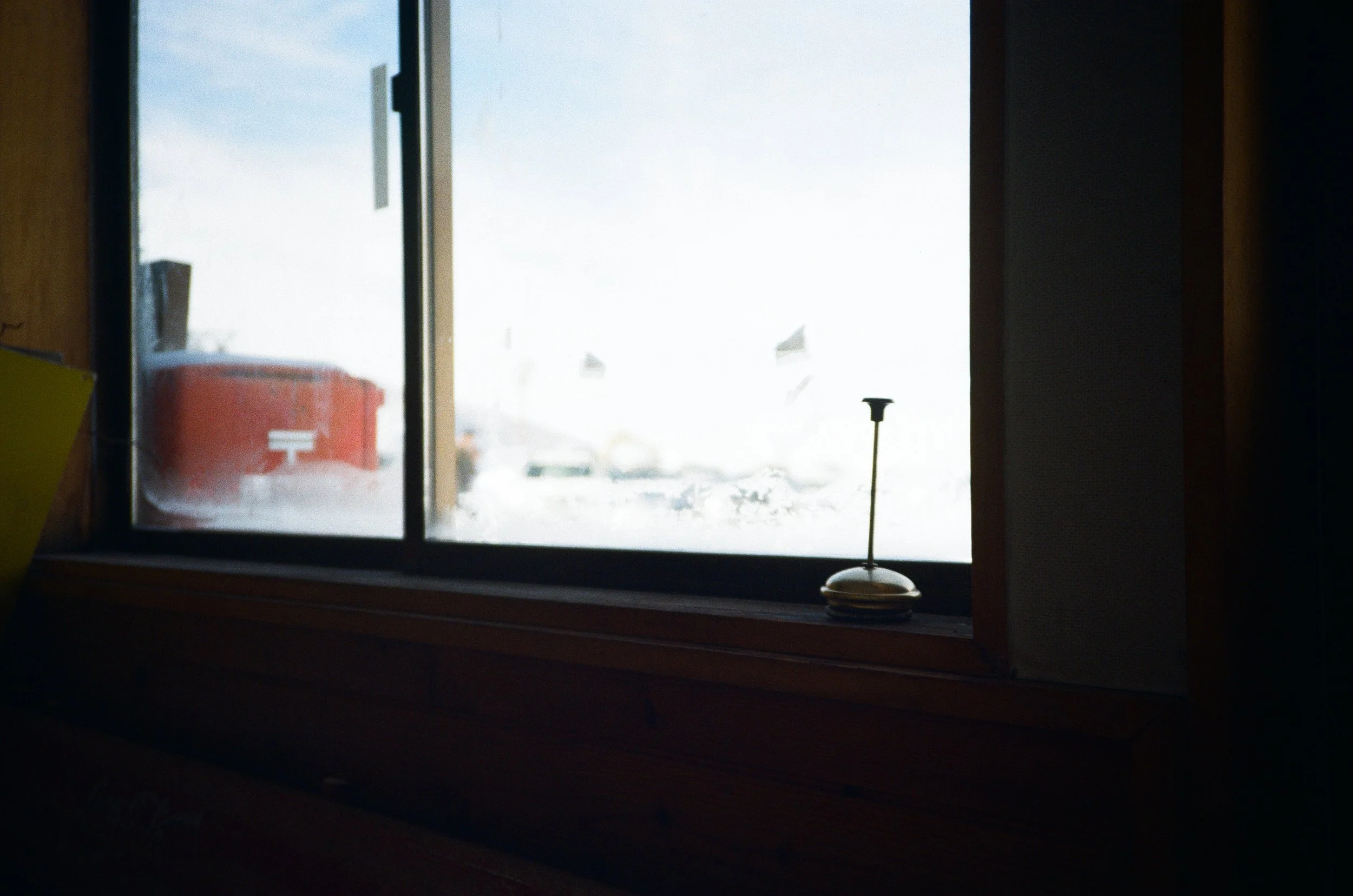 A view from inside a room looking out through a window on a snowy day. There is a small antenna placed on the windowsill. Outside, a red mailbox and some cars can be seen in the snow.