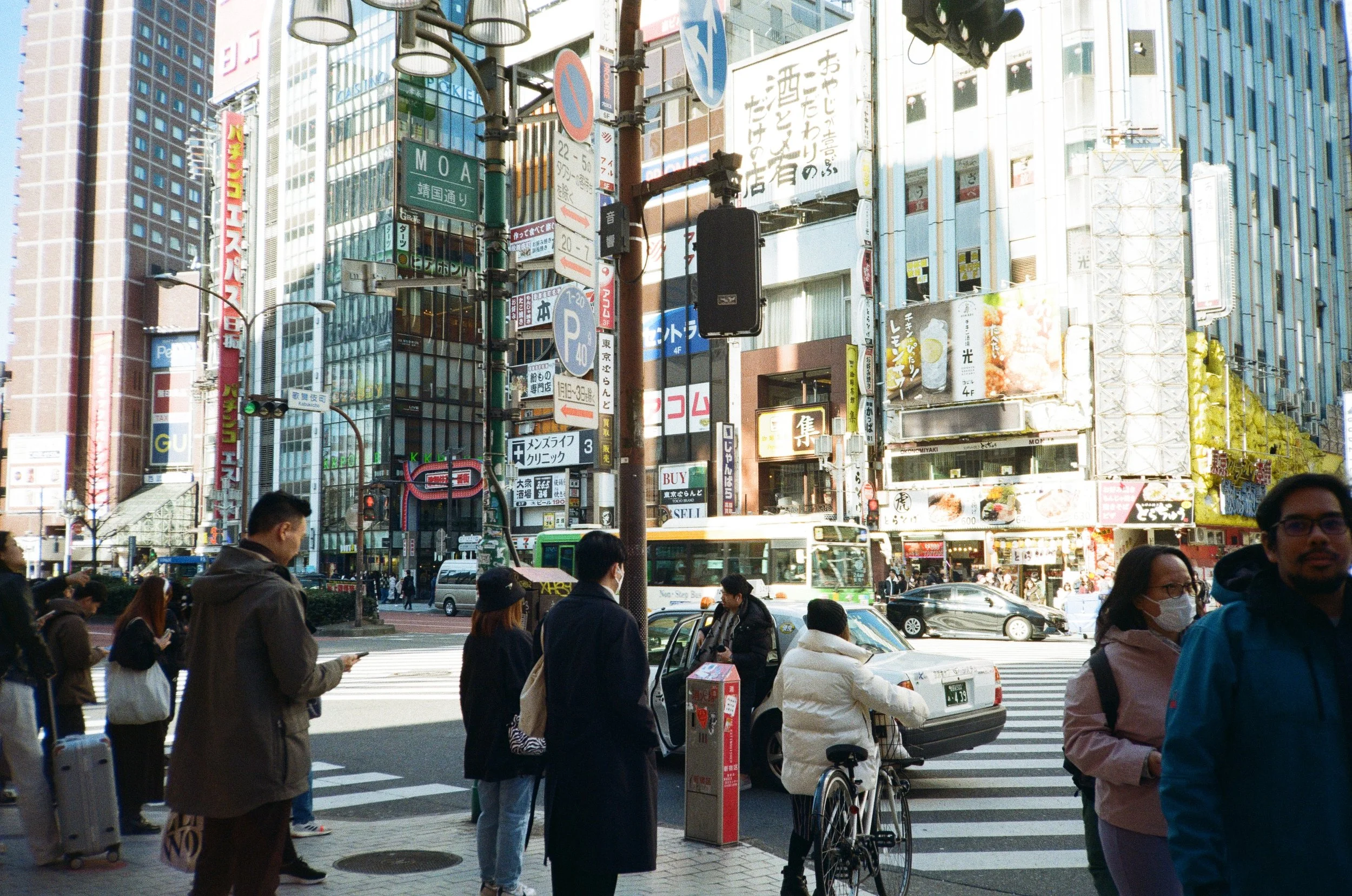 Crowd of pedestrians waiting at a crosswalk on a busy city street with tall buildings, billboards, and signs, some with Japanese characters, indicating an urban area in Japan.