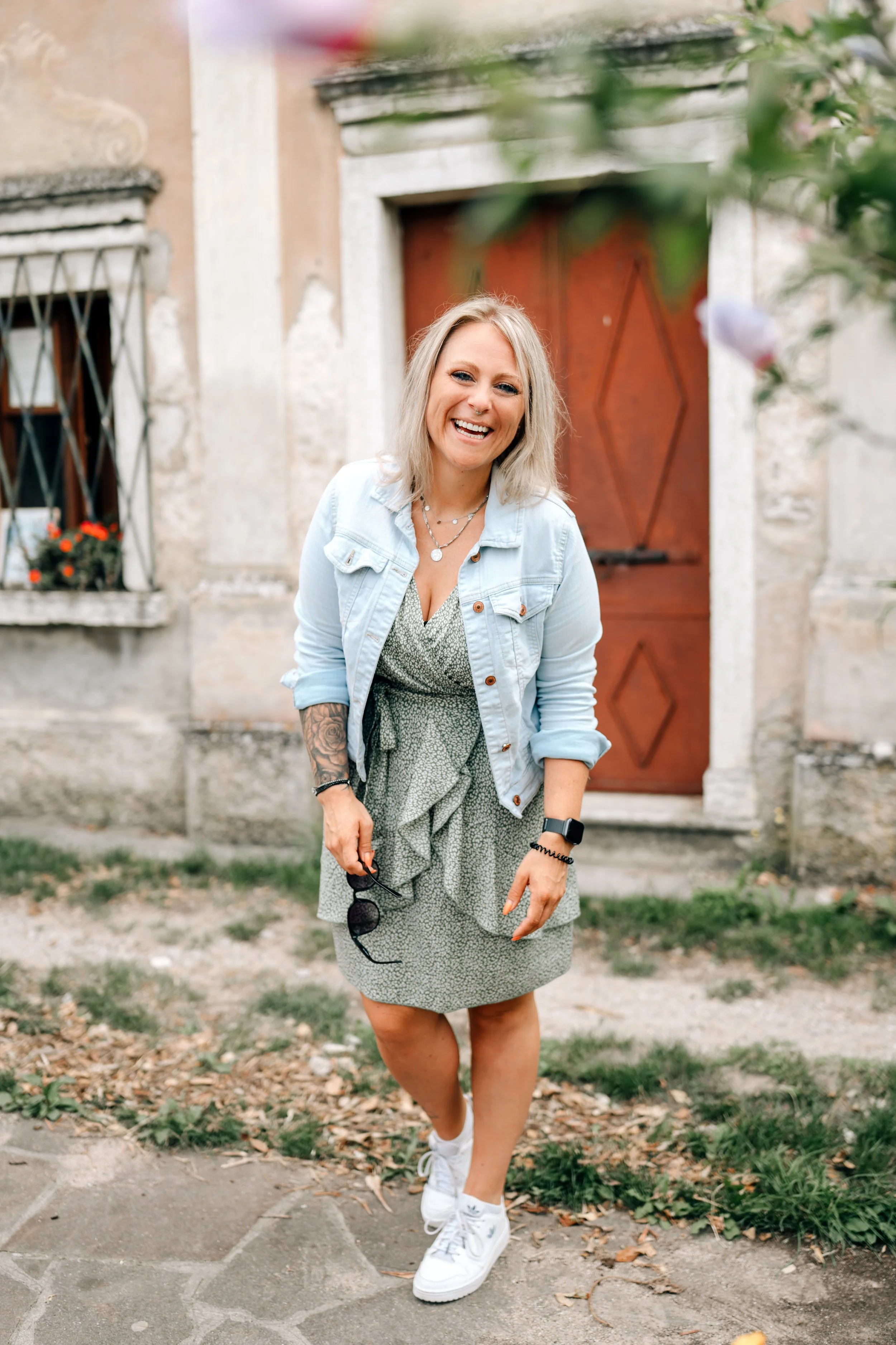 Eine lachende Frau mit blonden, mittellangen Haaren, trägt eine hellblaue Jeansjacke, ein grünes Kleid mit weißen Punkten, weiße Nike-Sneaker und hält eine Sonnenbrille in der Hand. Sie steht vor einer alten Steinmauer mit einem Holz-Tor und einer Fensterbank mit Blumen, im Garten auf einem gepflasterten Weg.